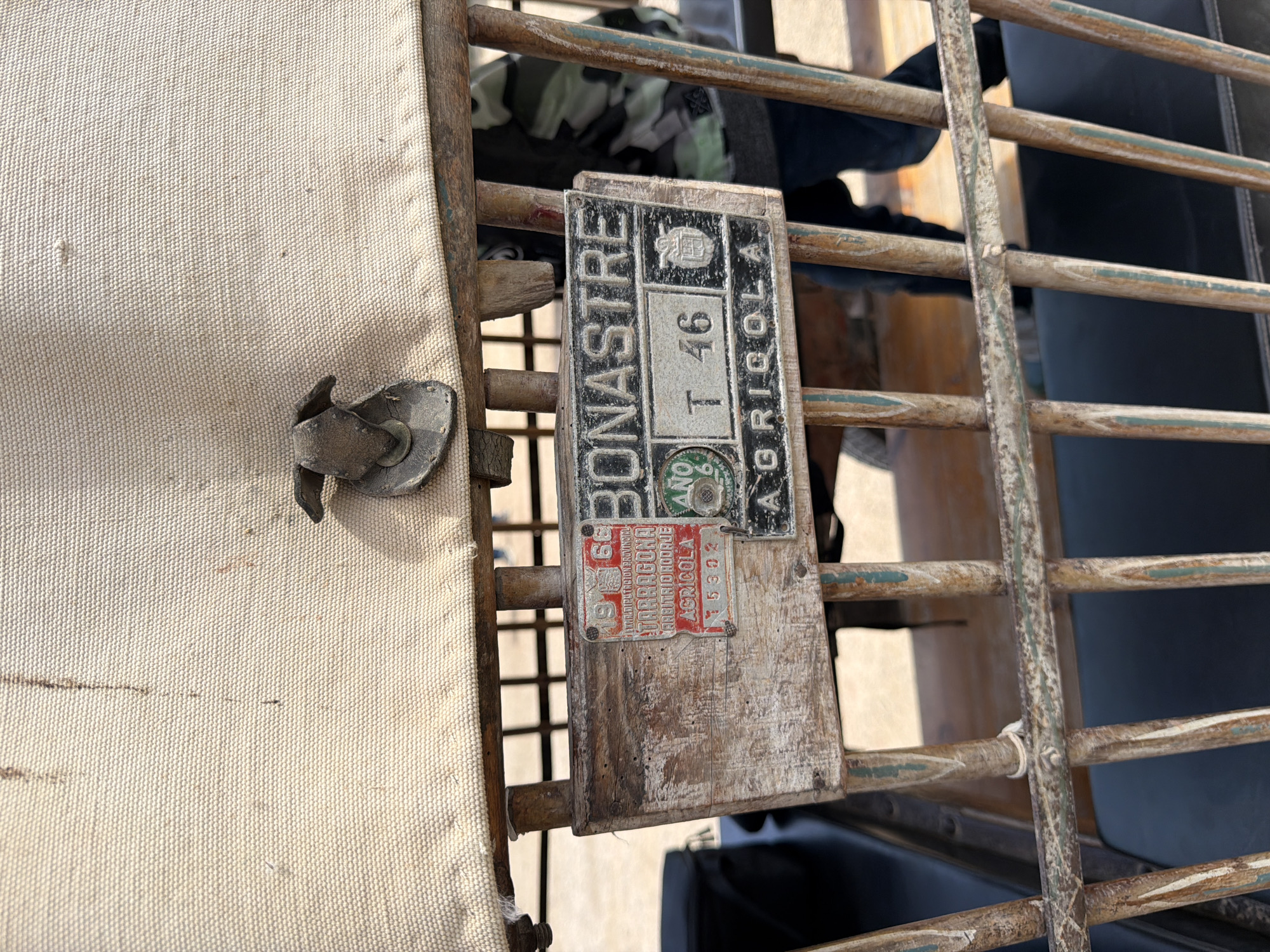 The image captures a close-up view of an aged agricultural registration plate and a fabric surface, likely part of a vehicle or cart. On the left, a coarse, off-white woven fabric, possibly canvas, is visible with a metal fastener or bracket attached. The fabric shows minor wear and some dark streaks.
The central subject is a rectangular metal plate, affixed to a weathered wooden board which is part of a metal-barred structure. This plate features "BONASTRE" written vertically on its left side. At the top, there's an emblem or coat of arms. Below this, "T 46" is prominently displayed. "AGRICOLA" is written vertically on the right side. A small green circular seal at the bottom of this plate clearly reads "AÑO 66", indicating the year 1966.
Below the main plate, a smaller, red rectangular plate is attached. It bears the text "19 G.C.", "EXCMO. PATRONATO PROVINCIAL", "TARRAGONA", "ARBITRAJE AGRICOLA", and the number "15302". This text, particularly "Bonastre", "Tarragona", and "Agrícola", strongly suggests the object is an agricultural vehicle or equipment registered in Bonastre, a municipality within the province of Tarragona, Spain, dating back to 1966.
Behind the wooden board and metal bars, glimpses of clothing, including camouflage fabric, are visible. The scene is illuminated by natural daylight, suggesting it was taken outdoors. There is no human activity present; the image is a still life focusing on the weathered objects and their historical context.