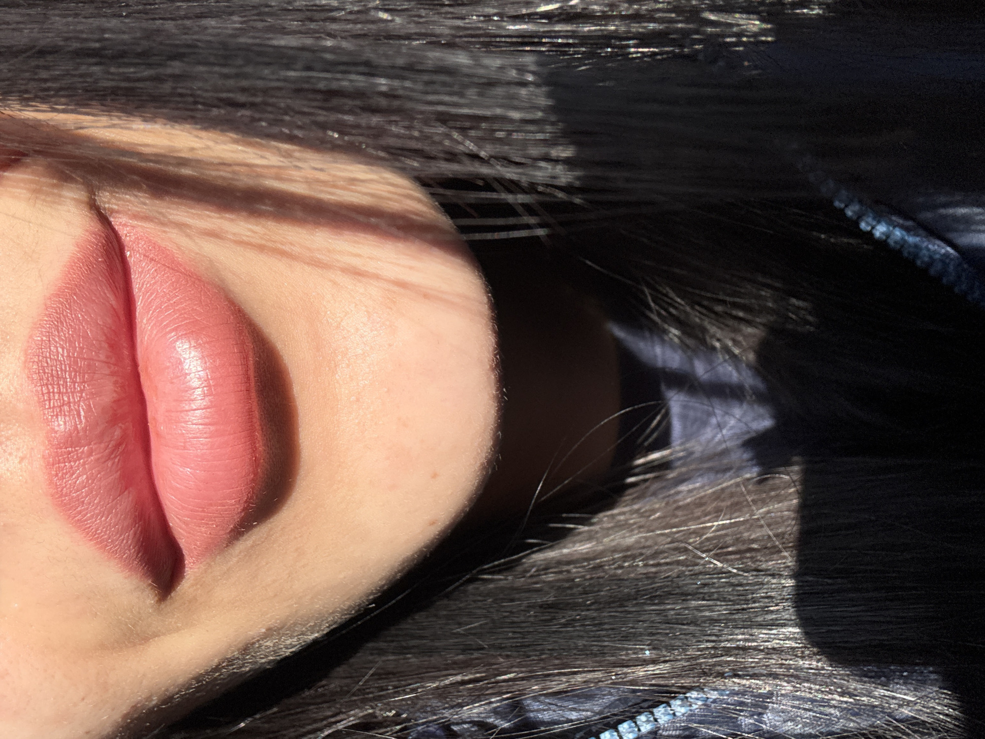 The image shows a close-up of a person's face, specifically focusing on the mouth and surrounding skin. The lips are painted with a pink lipstick, and the skin has visible pores and fine hairs. The hair is dark and appears to be lying around the face. The lighting is uneven, creating strong shadows on the face and hair. The presence of sequins on the clothing suggests a formal setting, and the scene location is Molodezhnyi, Russia.
