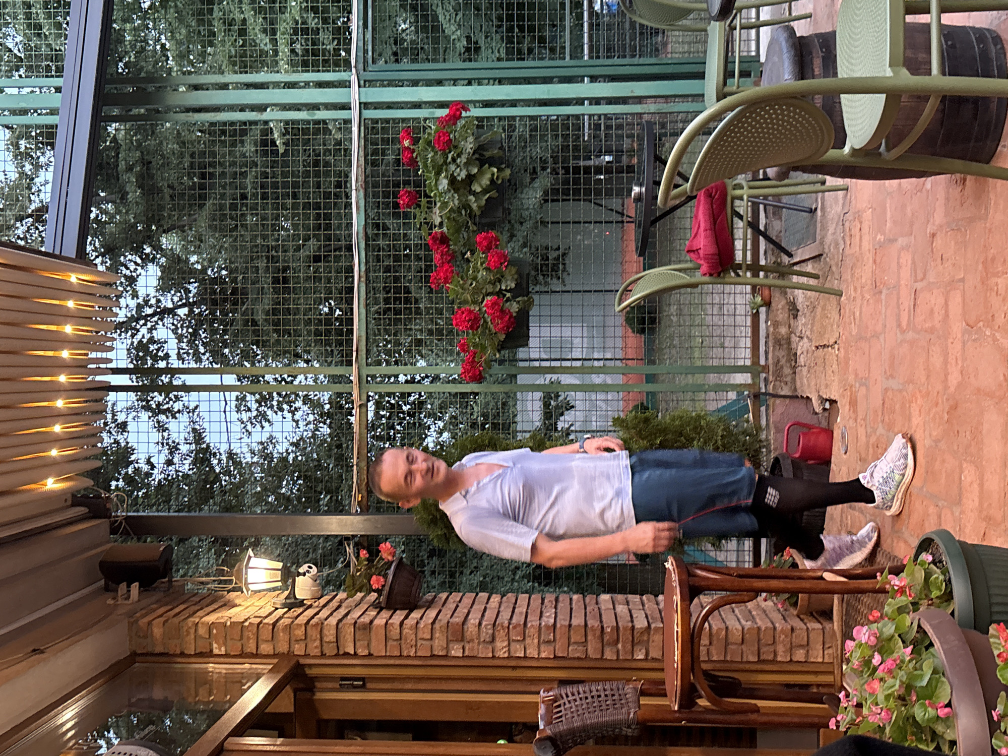 This image depicts an outdoor patio area in Cukarica Urban Municipality, Serbia, illuminated by artificial light.
One adult male is present, standing slightly left of the image center, facing partially right. He appears to be Caucasian, approximately in his 40s or 50s, with short, light brown hair. He is wearing a light blue short-sleeved collared shirt, dark blue shorts with a red vertical stripe on the outer seam, black compression socks extending to just below the knee, and multicolored athletic shoes (predominantly white, purple, and light green). His eyes are partially closed, suggesting a smile or squint.
The patio floor is composed of terracotta-colored paving tiles. To the far left, a brick column abuts a wooden doorframe and partially open door, suggesting an entrance to an indoor establishment. A dark-colored lantern with a warm glow is mounted on the brick column, with a small white security camera positioned directly below it. Two dark-colored planters containing pink/red flowers are attached to the brick column. A tall, dark wood bar stool with a woven seat and a small round wooden table are situated to the left of the male figure, near the brick column. In the bottom left foreground, a large dark planter contains green foliage and pink flowers.
The background is defined by a tall, dark green metal fence with a square grid pattern, supported by vertical green metal posts. Dense green trees are visible behind the fence. Two black hanging baskets with vibrant red geraniums are suspended from the metal fence. A small, conical evergreen shrub is planted on the ground directly behind the male figure, next to the fence. A red plastic watering can is visible on the ground behind the male's right foot, near the fence.
Multiple light olive-green metal chairs with perforated backs are arranged on the patio. One chair, positioned in the mid-ground, has a folded red garment on its seat. Two round, dark-colored metal tables are visible in the right half of the image; one has an ashtray. A dark wooden barrel serves as a base for one of the chairs on the far right. Overhead, a light-colored slatted awning or canopy is equipped with multiple embedded warm-toned lights, casting a glow on the immediate area.