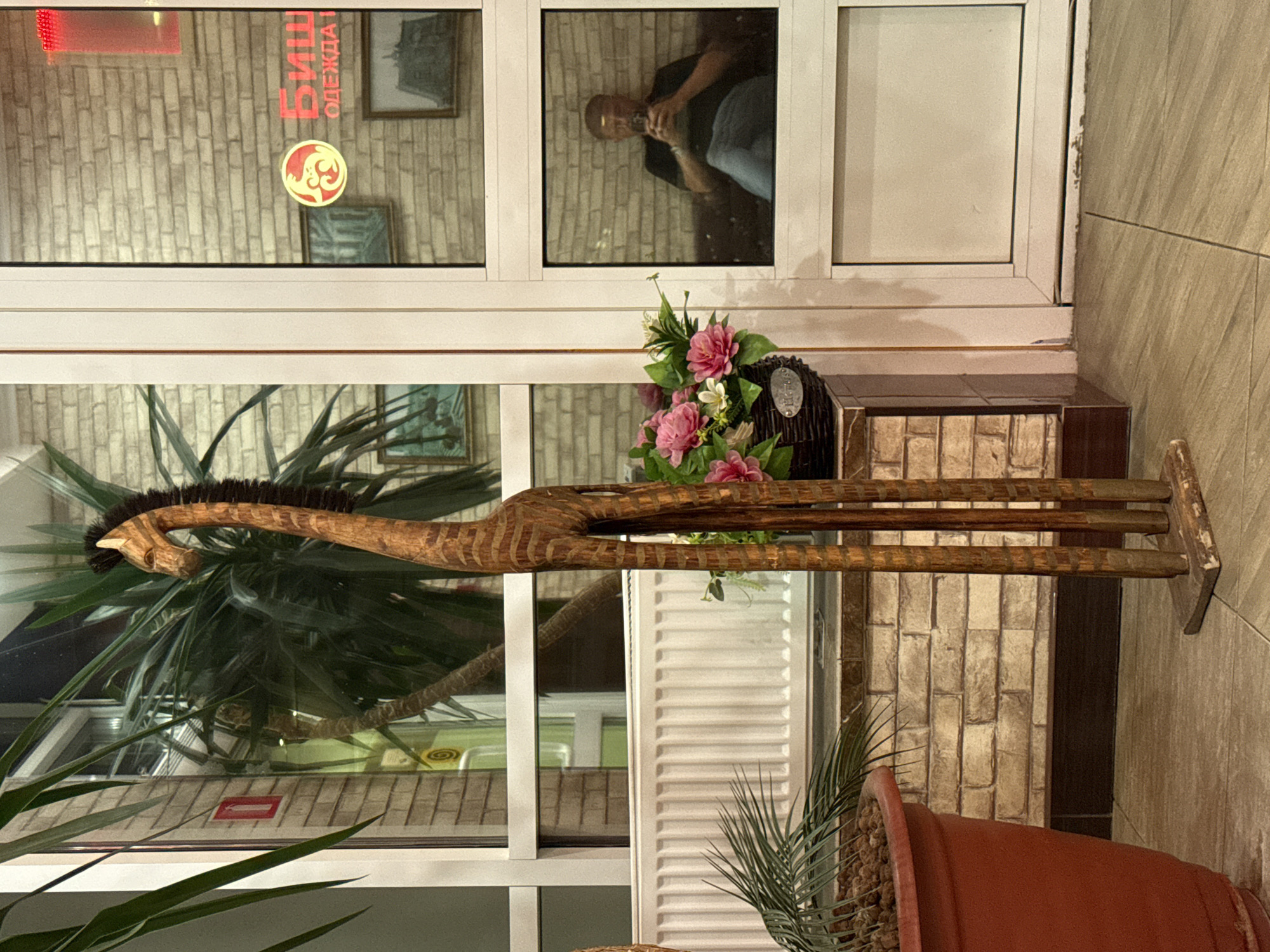 A wooden giraffe sculpture stands tall next to a large potted plant and a window with a reflection of a man using his phone. The setting appears to be an indoor space, possibly a shop or lobby, with brick-patterned walls visible through the window and a radiator behind the sculpture. The floor is tiled. The overall impression is one of a commercial or public indoor environment.