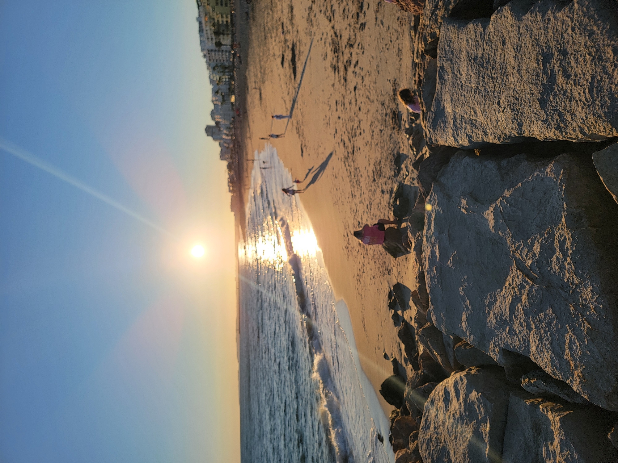 This is a vertically oriented photograph of a beach scene in Quarteira, Portugal, during sunset. The sun is low on the horizon, casting a warm, golden light across the sand and water, creating lens flare. The sky transitions from a clear blue on the left to a soft yellow and orange near the horizon.
The scene depicts a sandy beach with gentle waves rolling in from the ocean. A few people are scattered on the sand, some appearing to walk along the shoreline. One individual in the foreground is sitting on the sand, and another is visible further up the beach. Buildings line the distant edge of the beach, suggesting an urban or resort area. Large rocks, likely part of a seawall or jetty, dominate the right side of the frame, casting long shadows on the sand. The overall mood is peaceful and serene, characteristic of a sunset by the sea. There is no visible text in the image.