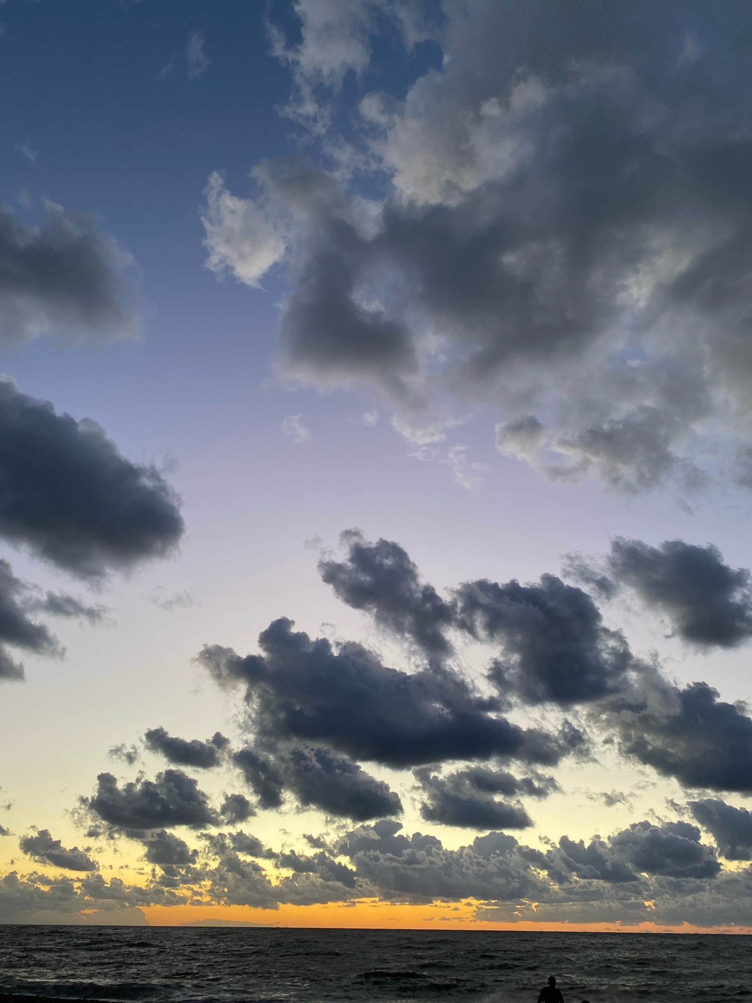 A dramatic sunset over a body of water is captured in this vertical shot. The sky transitions from a pale yellow and orange near the horizon to a soft lavender and then a deep blue at the top. Dark, heavy clouds are scattered across the sky, creating a stark contrast with the lighter parts of the sky. The water is dark and choppy, with visible waves. A silhouetted figure of a person is visible in the lower right corner, standing near the water's edge. The scene appears to be a coastal or lakeside environment.