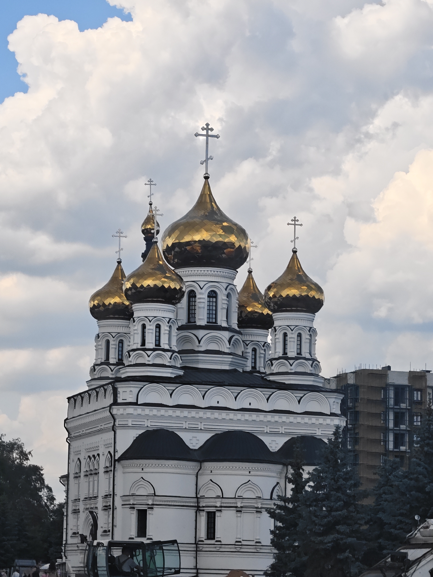 The image shows the Ascension Cathedral in Tver, Russia, on a partly cloudy day. The cathedral is a white building with multiple golden domes, each topped with a cross. The architecture is intricate, with arched windows and decorative moldings. In the foreground, dark green coniferous trees obscure the lower part of the cathedral. To the right of the cathedral, a modern building under construction, covered in scaffolding, is visible. A portion of a vehicle with a visible driver and a person in the passenger seat is in the bottom center of the frame, suggesting ongoing activity around the site. The sky is a mix of bright blue patches and large, fluffy white and gray clouds.