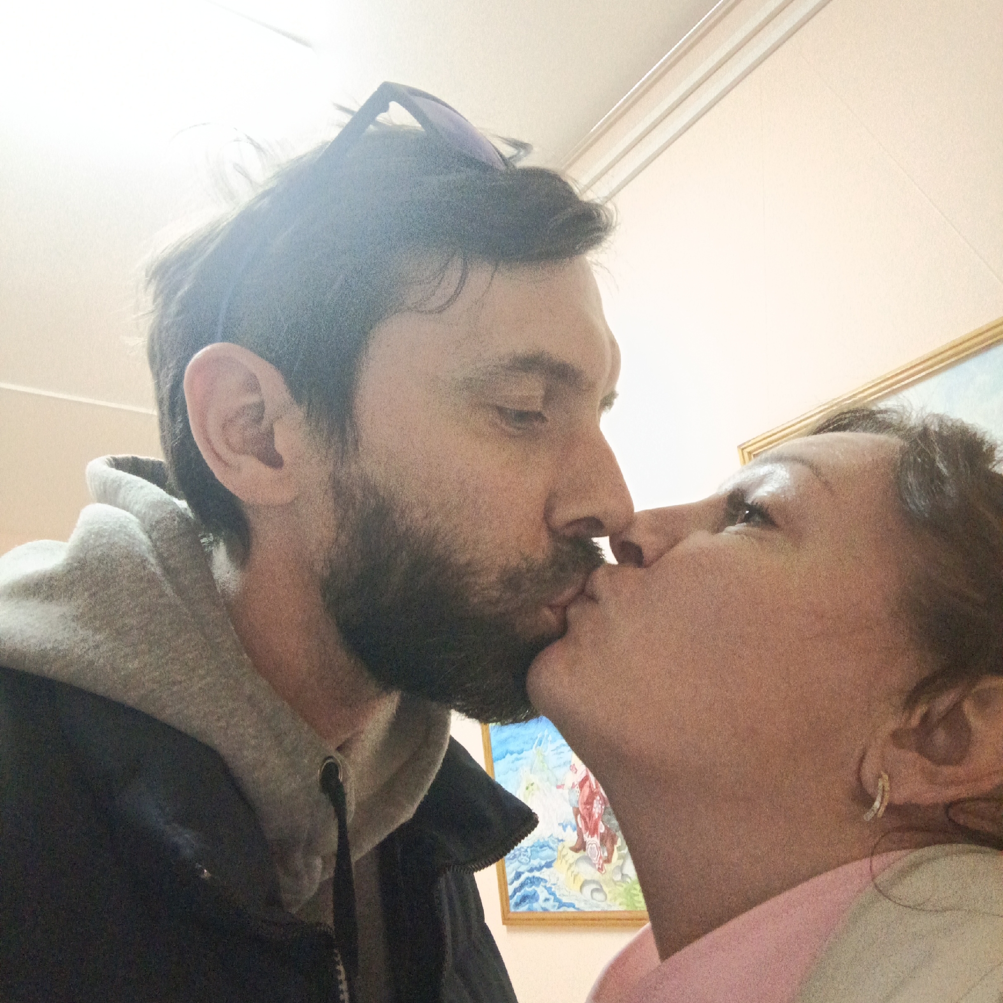 A close-up photograph shows a man and a woman kissing. The man has a dark beard and is wearing a gray hoodie under a dark jacket. Sunglasses are perched on his head. The woman is wearing a pink shirt and has hoop earrings. In the background, part of a framed painting is visible. The location appears to be indoors, potentially in Markovskii, Russia.