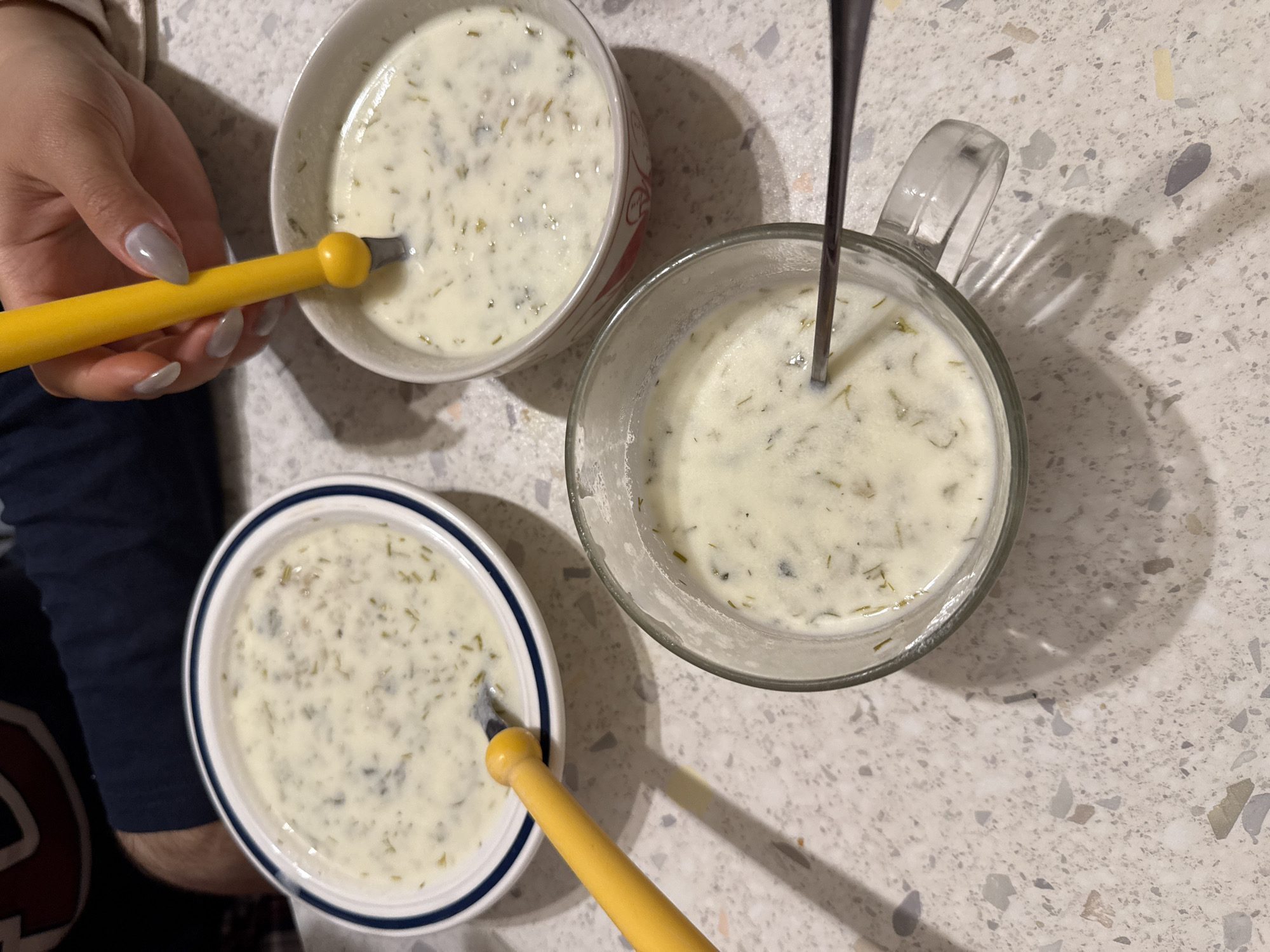 The image presents a top-down view of three servings of a creamy soup, likely cacik, set on a speckled countertop. One serving is in a white bowl, another in a blue-rimmed bowl, and the third in a glass mug. Two of the servings have yellow-handled spoons, and the third has a metal spoon. A person's hand is visible near the white bowl. The location is not identifiable as Milan, Italy from the visual information provided.