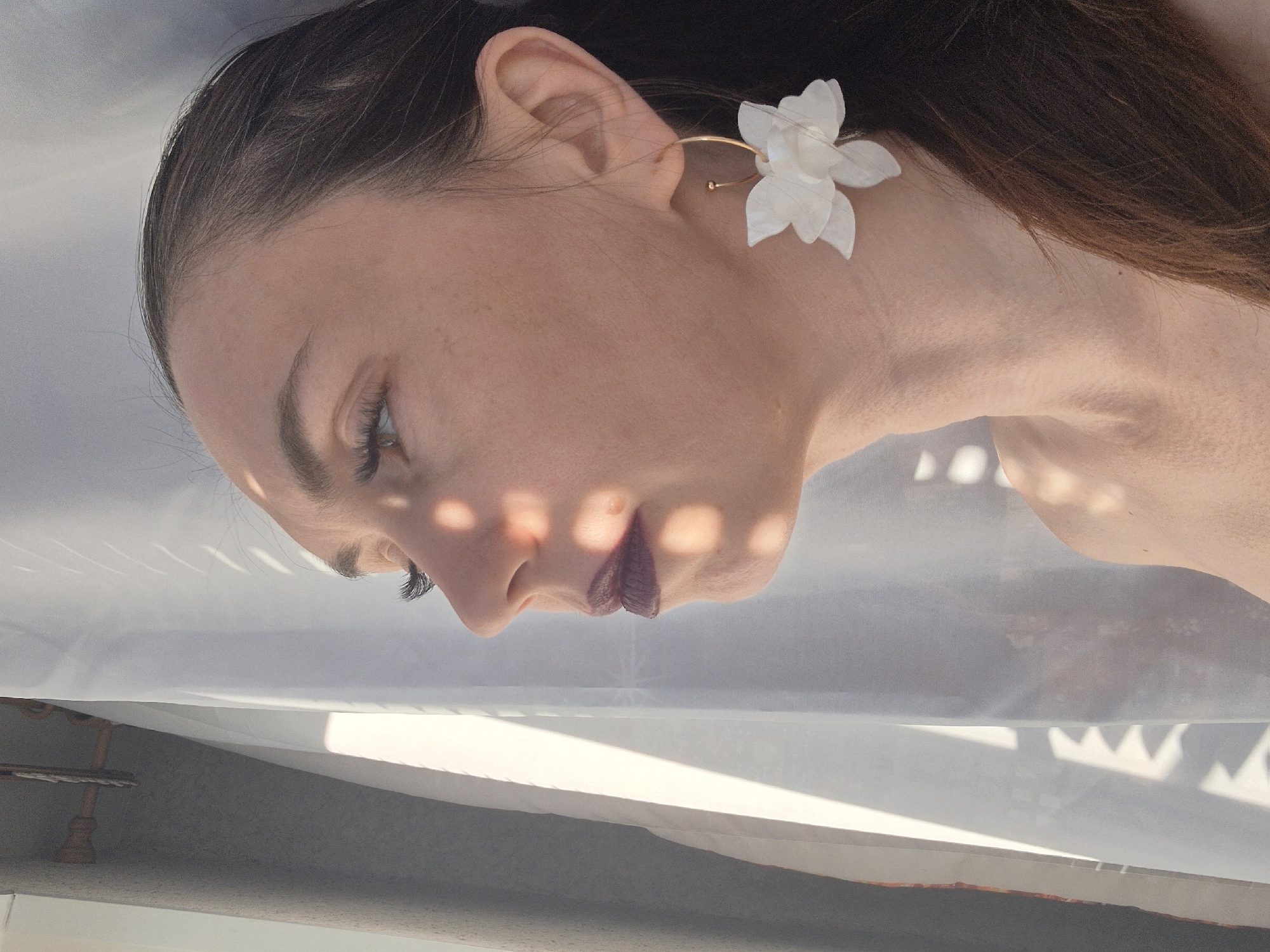 A close-up portrait shows a person's face in profile, angled downward. The person has fair skin, dark hair pulled back, visible eyelashes, and lips colored with a dark shade. They are wearing a gold hoop earring adorned with a white flower. The person is partially obscured by a translucent fabric, possibly a curtain, which casts dappled light across their face. Below the fabric, there is a view of a gray surface, possibly a floor or table, with part of a light-colored wooden object visible at the bottom left. The background city and location are unknown.