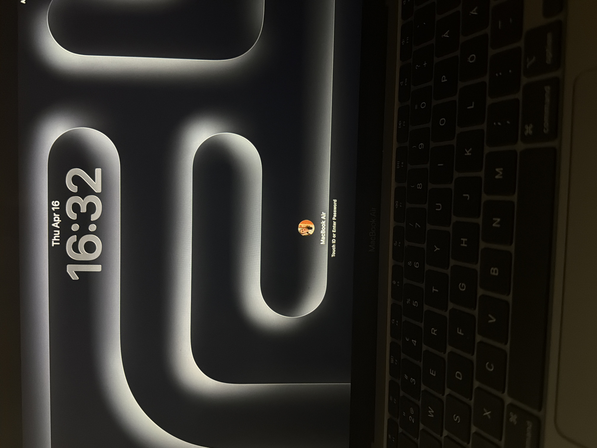 An overhead, slightly angled shot captures a MacBook Air laptop at its lock screen, set in an indoor environment in Plovdiv, Bulgaria. The laptop's screen occupies the left side of the frame, displaying a dark background with illuminated, abstract, rounded white and grey shapes. Prominently featured on the left of the screen are the date and time, "Thu Apr 16" and "16:32". To the right of the screen, a small circular profile picture of a user is visible, along with the device name "MacBook Air" and the login prompt "Touch ID or Enter Password".
The right half of the image shows the laptop's keyboard, which is dimly backlit, suggesting low ambient lighting conditions. The keys are discernable, featuring a standard QWERTY layout, but also displaying characters such as 'Å' and 'Ö', indicating a Nordic or similar regional keyboard configuration. The image depicts the laptop in a state awaiting user authentication during the late afternoon.