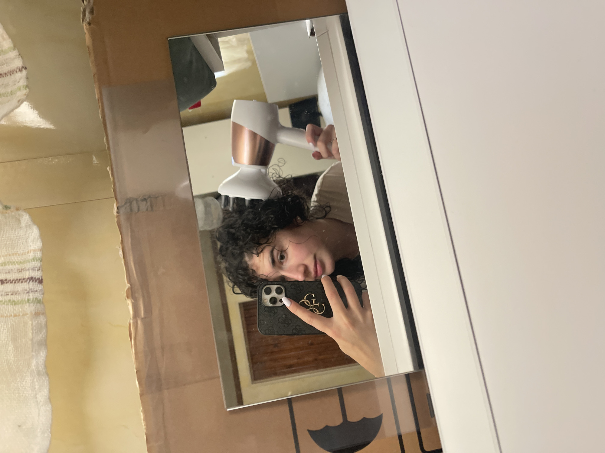 A person with curly dark hair is reflected in a mirror while holding a cell phone in their right hand and a white and rose gold hair dryer in their left. The phone has a dark-colored case with a gold emblem. The background in the mirror appears to be a bathroom. The mirror is mounted on a cardboard surface, which is next to a white cabinet or wall. A towel is visible to the left of the cardboard.
Given that there are no details present within the image that explicitly describe or identify the city of Sofia, Bulgaria, the location is unknown.
