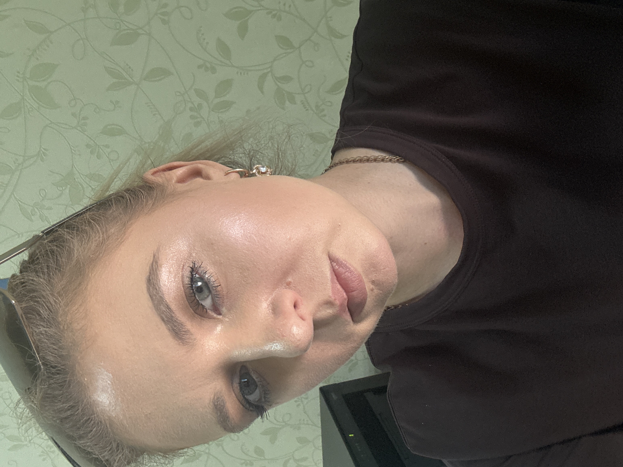 This is a close-up, slightly over-the-shoulder selfie of a person with blonde hair. The person is wearing a dark top and a gold chain necklace. Their face is turned towards the camera, with one eye visible and looking at the viewer. Their skin appears smooth and well-lit, with some subtle makeup. They are wearing earrings and a ring on one hand. The background consists of a light green wallpaper with a subtle floral pattern. The image is oriented sideways.