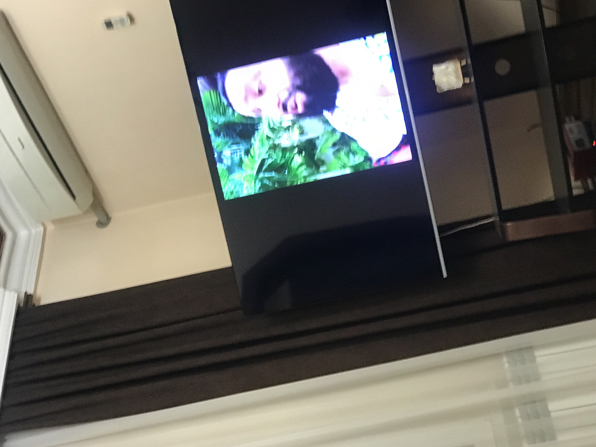 A television displays a human figure in a garden scene. An air conditioning unit is mounted on the wall. The setting is an interior space with dark brown horizontal blinds. The city and country are Yerevan, Armenia. The description is based on information in the image.