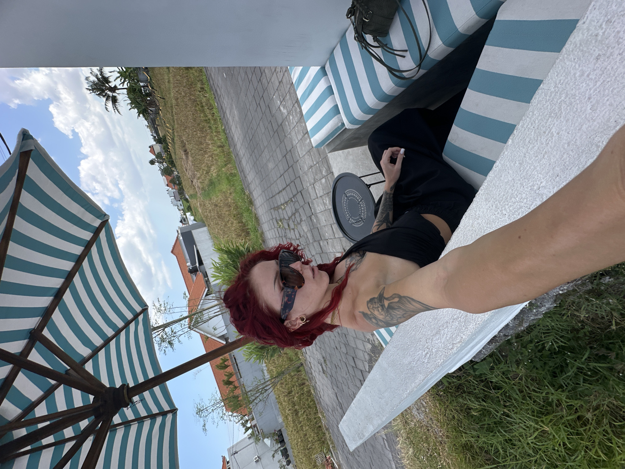 A woman with bright red hair and tattoos on her arm sits on an outdoor bench with blue and white striped cushions. She is wearing large, dark-rimmed sunglasses and a black top. To her left, a large umbrella with blue and white stripes is partially visible. In the background, there are buildings with orange tile roofs, palm trees, and a field of dried rice paddies, suggesting a location in the rural outskirts of North Kuta, Indonesia. A small black side table is positioned next to her, and a dark green handbag rests on the bench. The ground is paved with gray interlocking stones. The overall scene depicts a relaxed, outdoor setting under a partly cloudy sky.