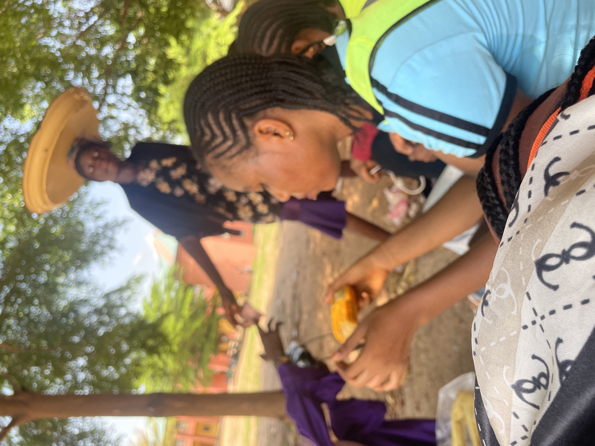 An outdoor scene, likely in Lokoja, Nigeria, depicts several young women and a vendor. The foreground features a woman in a light blue shirt with black stripes, wearing a bright yellow vest. Her hands are visible, holding a piece of food. She has braided hair. Beside her, another person's braided hair is also visible. In the middle ground, a vendor with a large, flat, yellow tray balanced on her head, wears a dark jacket over a patterned dress. She is interacting with another person whose hands are reaching out, possibly for a transaction. Another individual, partially obscured, wears a purple garment. The background shows trees with dappled sunlight, suggesting a park or open area, and a building with reddish-orange walls. The overall impression is a lively, everyday interaction in an urban environment.