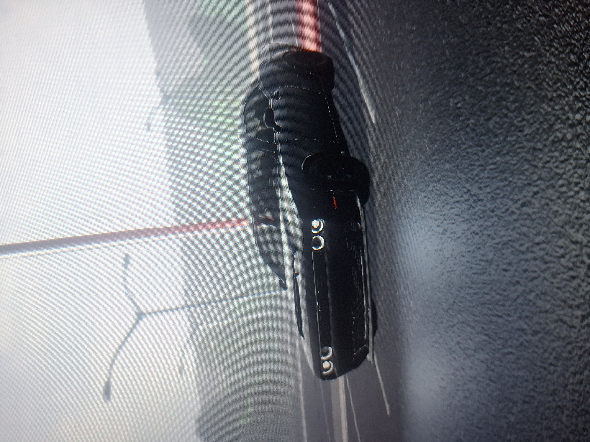 The image depicts a black car on a wet asphalt road in an urban setting. Light posts are visible in the background. The location is possibly in Sofia, Bulgaria, based on the environment and architecture. No people are visible.
