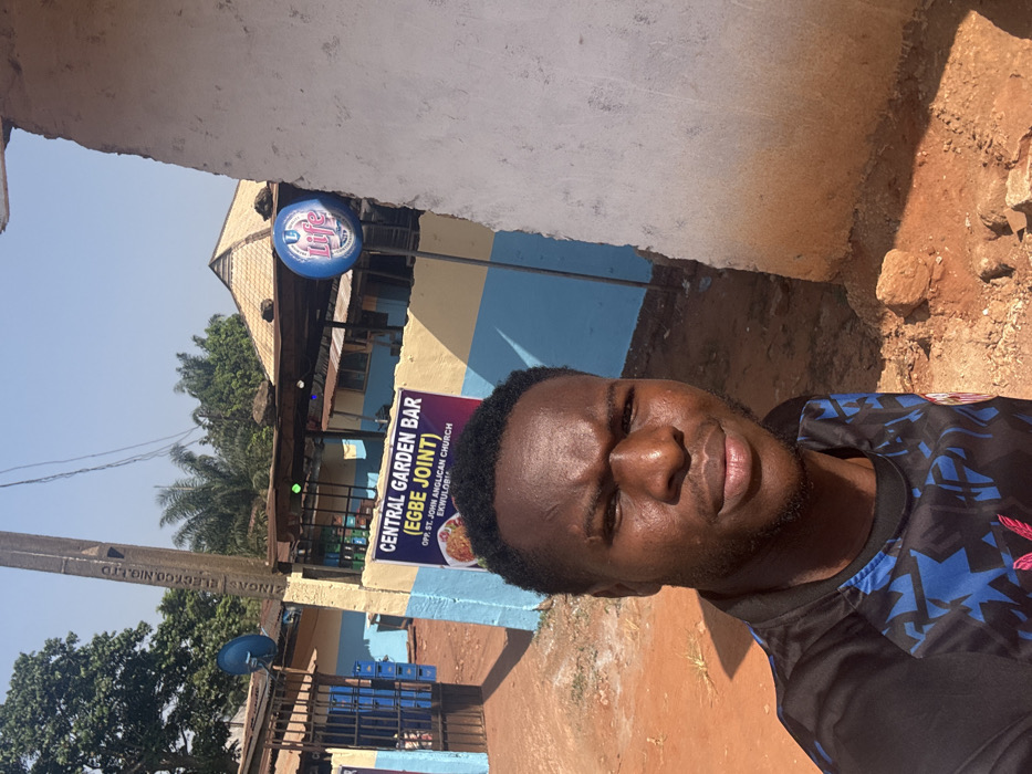 An outdoor scene in Ekwulobia, Aguata, Nigeria, depicts a dark-skinned adult male taking a selfie in the foreground. The male has short, dark hair, light facial stubble, and visible scars on his forehead, wearing a dark-patterned t-shirt with a red emblem. The setting is characterized by reddish-brown dirt ground under a clear blue sky.
In the midground, from left to right, a dark blue metal gate opens to a compound, with stacked blue plastic crates and a blue satellite dish visible near it. A utility pole marked "ZINGAS ELECTRO.NIG.LTD" stands prominently. Adjacent to this, a building with a light blue and yellow-cream painted lower facade features a large sign for "CENTRAL GARDEN BAR (EGBE JOINT)," specifying its location as "OPP. ST. JOHN ANGLICAN CHURCH EKWULOBIA" and displaying a food image. To the right of this structure, a blue circular "Life" beer sign on a pole is positioned next to a partially visible plain light-colored concrete wall. The background includes green trees and vegetation, notably palm trees, and a distant building with a multi-tiered, conical roof structure. Strong daylight illuminates the scene, creating distinct shadows.