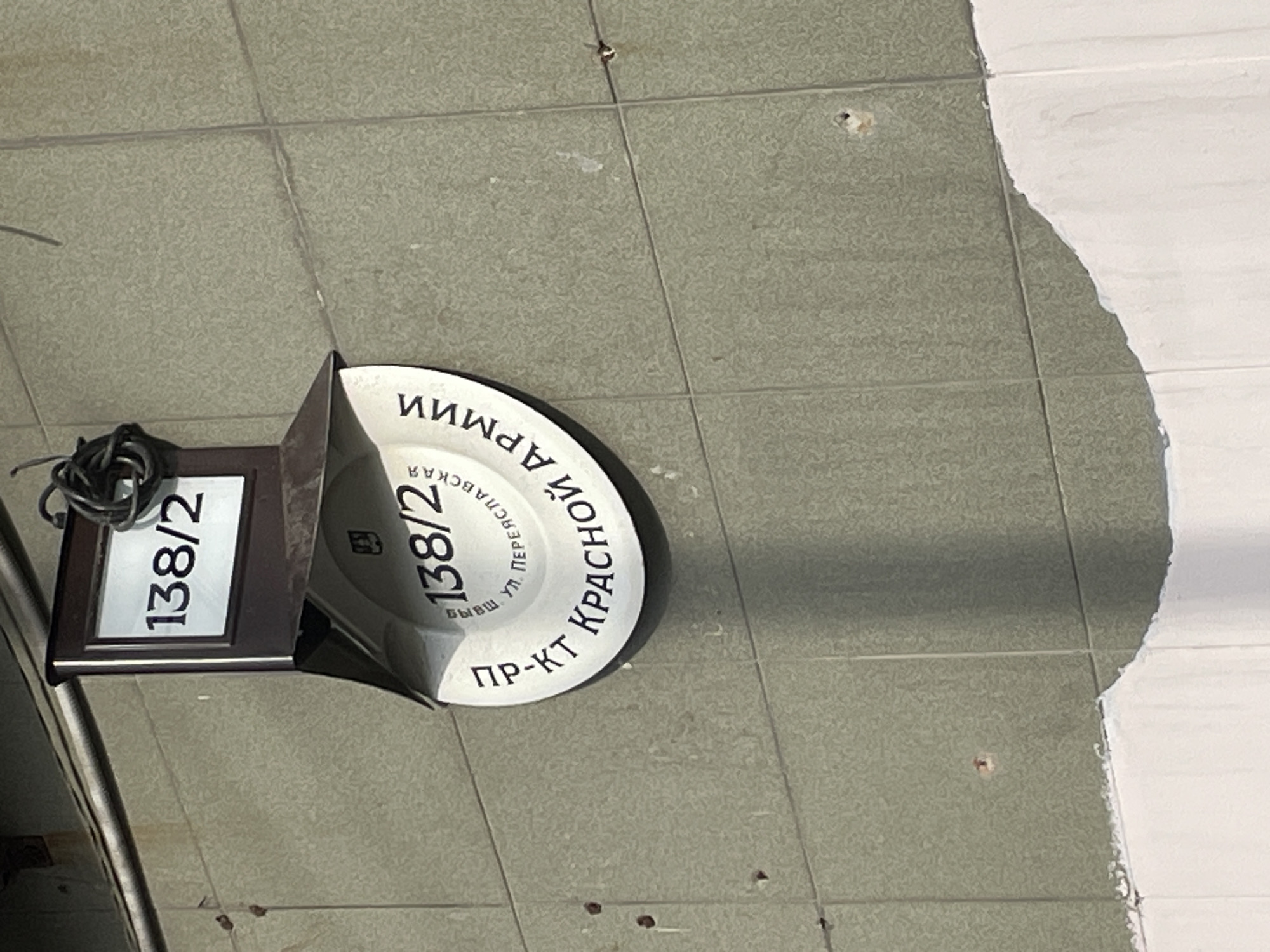 The image shows a building facade in Sergiyev Posad, Russia. A sign with the number "138/2" is mounted on the wall. Below this, a circular sign displays "138/2" and Russian text that translates to "pr-kt Krasnoy Armii" (Krasnaya Armiya Avenue) and "vyssh ul. Pereyaslavskaya" (higher street Pereyaslavskaya). The building is covered in green tiles, and there is a section of white plaster at the bottom of the image. The lighting suggests it is daytime, and the overall scene is an outdoor urban environment.