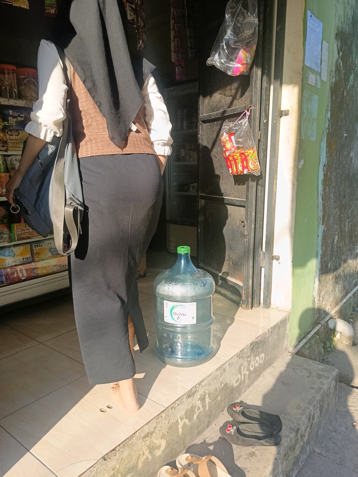 The image depicts a street view in Makassar, Indonesia, on a sunny day. The foreground shows a person from the waist down, wearing a dark grey, ribbed, floor-length skirt and a white long-sleeved shirt. The person is carrying a dark blue bag over their left shoulder and is barefoot, standing on a tiled surface. To the right, a large, clear plastic water jug with a green cap is positioned on the tiled ground. The background features the entrance to a small shop or kiosk with a dark metal door. Several plastic bags containing snacks or small items are hanging from the doorframe. Shelves with packaged goods are visible inside the shop. The wall to the right of the entrance is painted light green and appears weathered, with some graffiti or writing on the concrete step in front of it. A pair of dark grey sandals with red heart embellishments and a pair of tan sandals are placed on the concrete step. The lighting suggests it is daytime, with strong sunlight casting shadows.