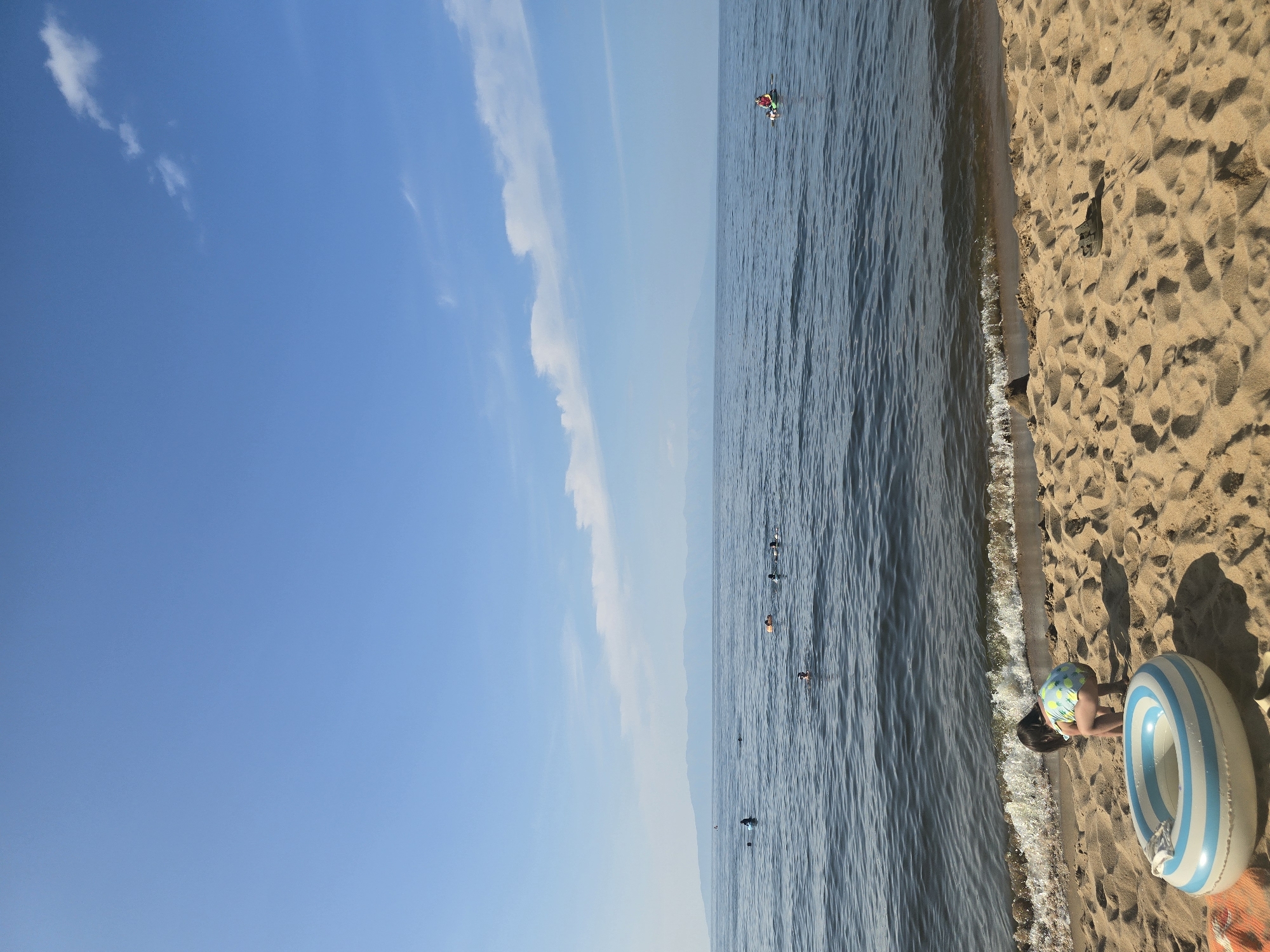 A daytime, vertical shot captures a serene beach scene in Maksimiha, Russia. The foreground is dominated by golden sand, scattered with shadows and footprints. A blue and white striped inflatable ring lies on the sand near the water's edge, beside a child wearing a pineapple-patterned swimsuit who is crouched down, facing away from the camera and towards the water. The water, a deep blue, gently laps onto the sandy shore with small waves.
Further out in the sea, several people are visible swimming or wading. One individual is on a small inflatable raft in the middle distance. In the background, a hazy, mountainous shoreline stretches across the horizon under a clear blue sky dotted with a few wispy white clouds. The overall impression is one of a peaceful summer day at the beach.