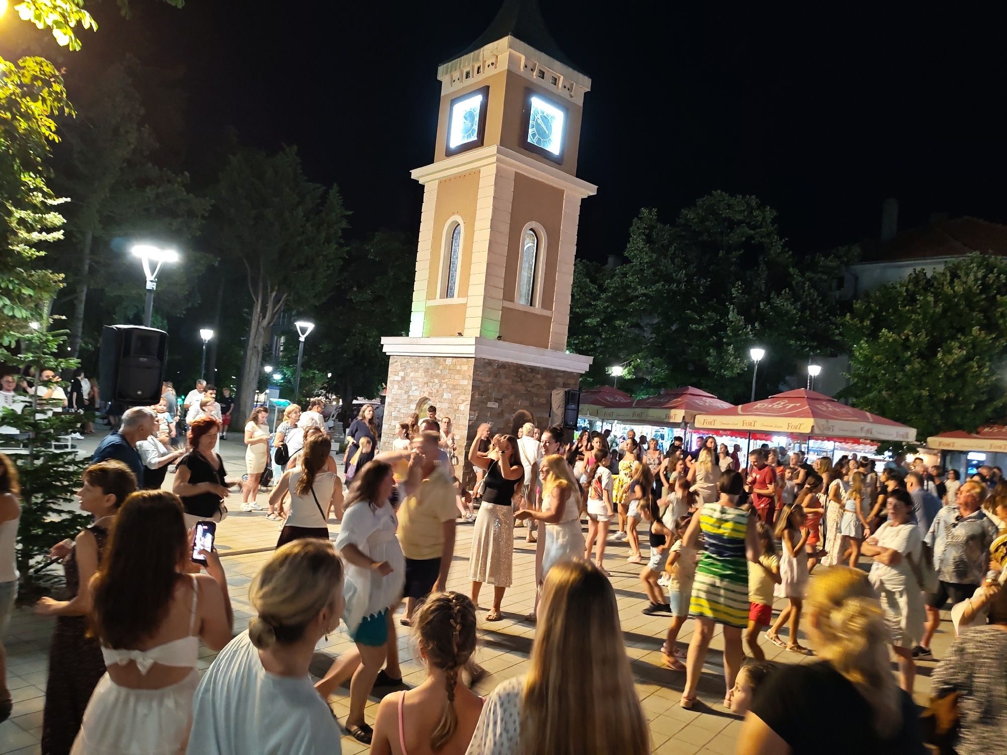 This is a nighttime photograph taken in the city of Obzor, Bulgaria, likely in the central square area, as indicated by the presence of a prominent clock tower. A large gathering of people is visible, appearing to be engaged in a festive event or celebration, possibly dancing. The clock tower, illuminated and displaying the time, serves as a central focal point. Surrounding the tower and the crowd are trees, streetlights, and structures that resemble outdoor seating areas or market stalls, some covered with red awnings bearing the logo "Fort". The ground is paved with cobblestones or similar material. The overall atmosphere suggests a lively public event taking place after dark.