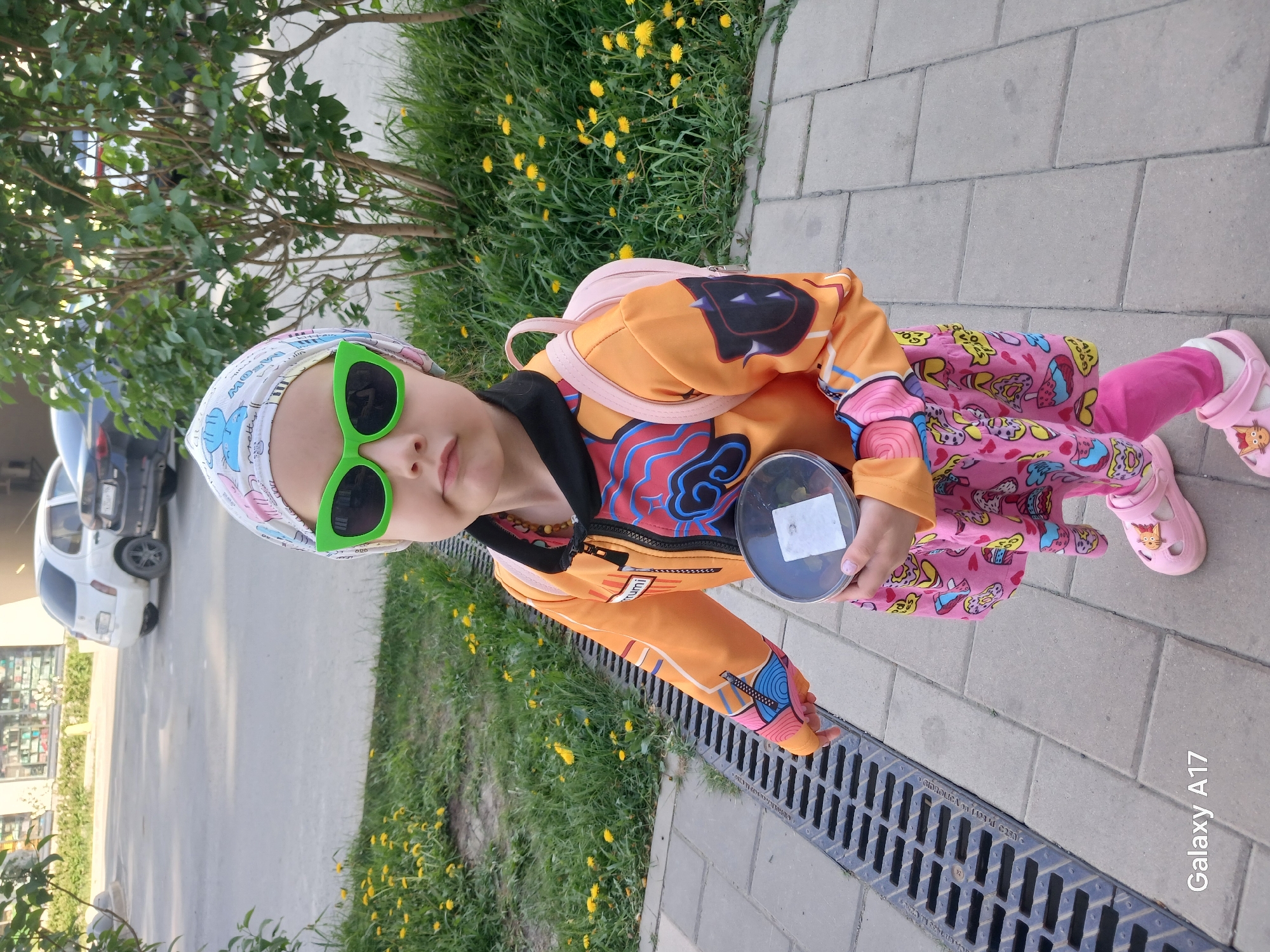 A young child is standing outdoors. The child is wearing green sunglasses, a patterned headscarf, an orange jacket with colorful designs, a pink patterned dress, and pink leggings. They are also wearing pink shoes with cartoon characters. The child is holding a small container. The environment consists of a paved sidewalk, grass, and yellow flowers. A tree is visible in the background, along with parked cars and a building. A drain is near the child. The "Galaxy A17" watermark indicates the image was taken with a Samsung Galaxy A17. The location is Yekaterinburg, Russia.