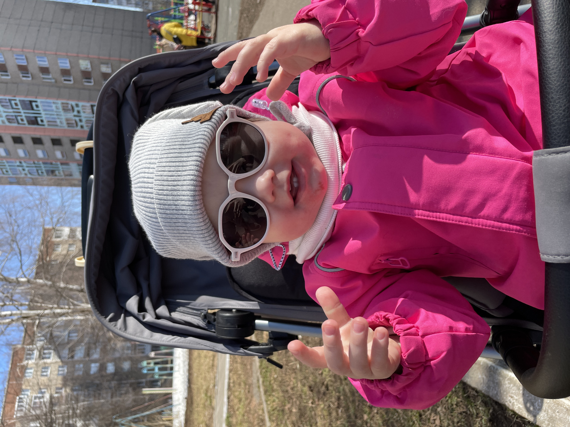 A young child wearing a gray knitted hat and white-framed sunglasses sits in a gray stroller. The child wears a bright pink jacket and holds their hands up. The setting is outdoors, possibly a park or playground, with a building visible in the background. The location is Izhevsk, Russia.