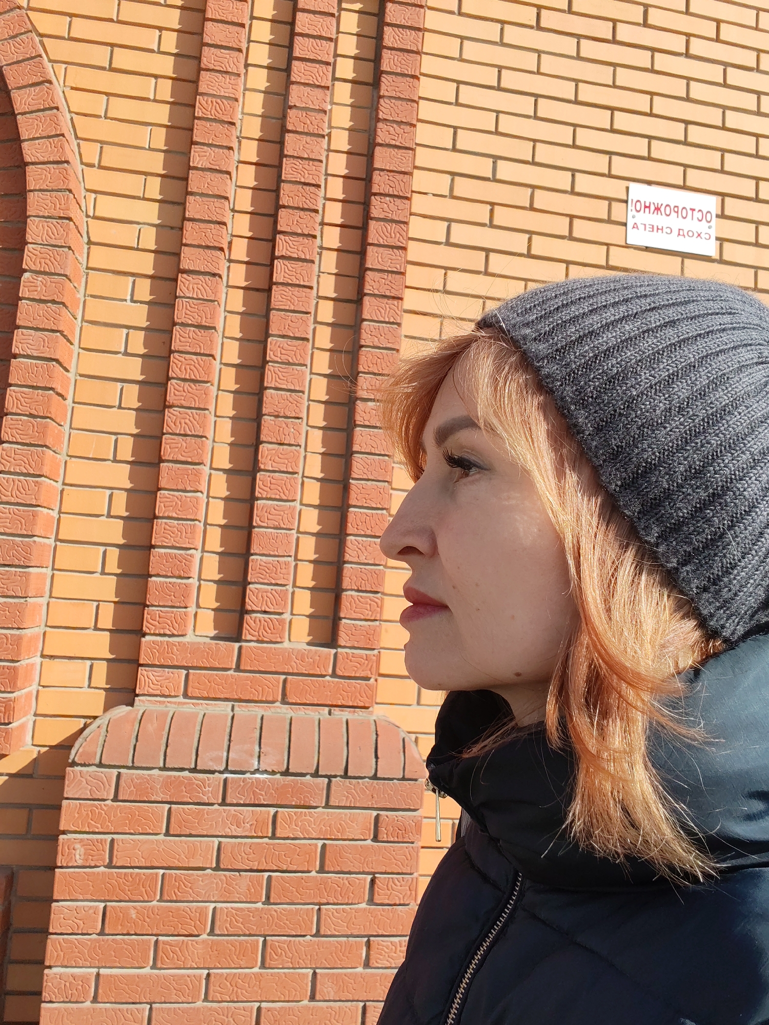 A woman is depicted in a close-up, right-profile view, looking to the left of the frame. She has reddish-blonde hair partially covered by a dark grey knitted beanie. She wears a black puffer jacket with a visible zipper pull on the right side of her neck. Her right eye shows visible eyeliner and mascara, and her lips appear to have a reddish tint. A small, dark mole is present on her right jawline. The background consists of a brick wall constructed from two types of bricks: lighter, smoother orange bricks forming the primary wall, and darker, reddish-brown bricks with a textured, wavy surface arranged in vertical decorative columns and a partial archway on the far left. Strong sunlight casts sharp shadows from the brickwork. A white rectangular sign with red inverted Cyrillic text, "ОСТОРОЖНО! СХОД СНЕГА" (CAUTION! SNOW SLIDE), is affixed to the upper right portion of the brick wall. The scene is situated outdoors in the city of Chaikovsky, Russia.