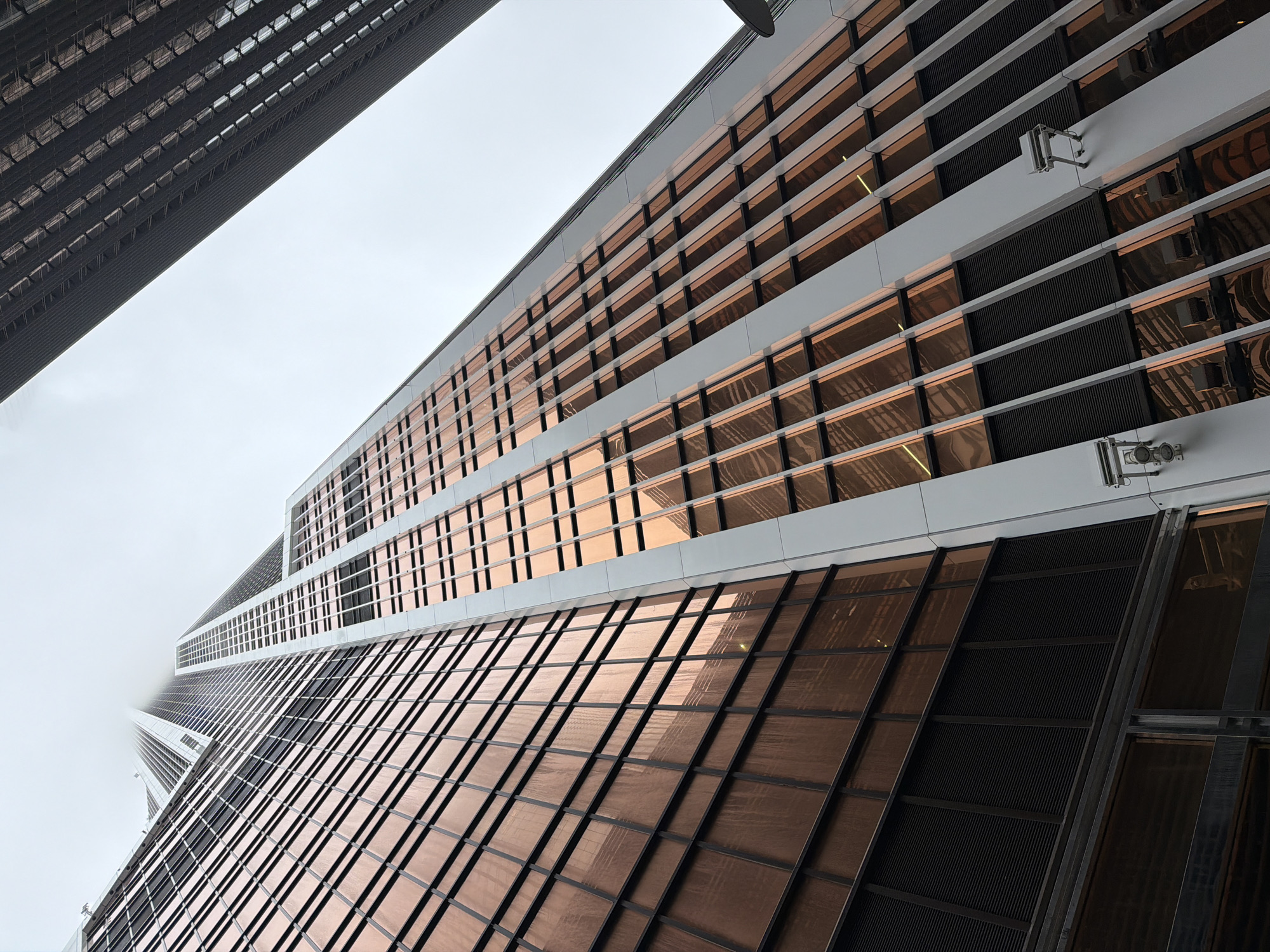 The image shows a low-angle view of skyscrapers in Moscow, Russia. The architecture features a modern design with numerous glass windows and metal accents. The building on the right is characterized by horizontal layers of windows and dark panels, with some metallic fixtures attached to the facade. In the upper-left corner, the top of another skyscraper is visible, its surface densely covered with small, square windows. The sky is overcast and obscured by fog, particularly in the distance where the skyscrapers appear to merge with the clouds.