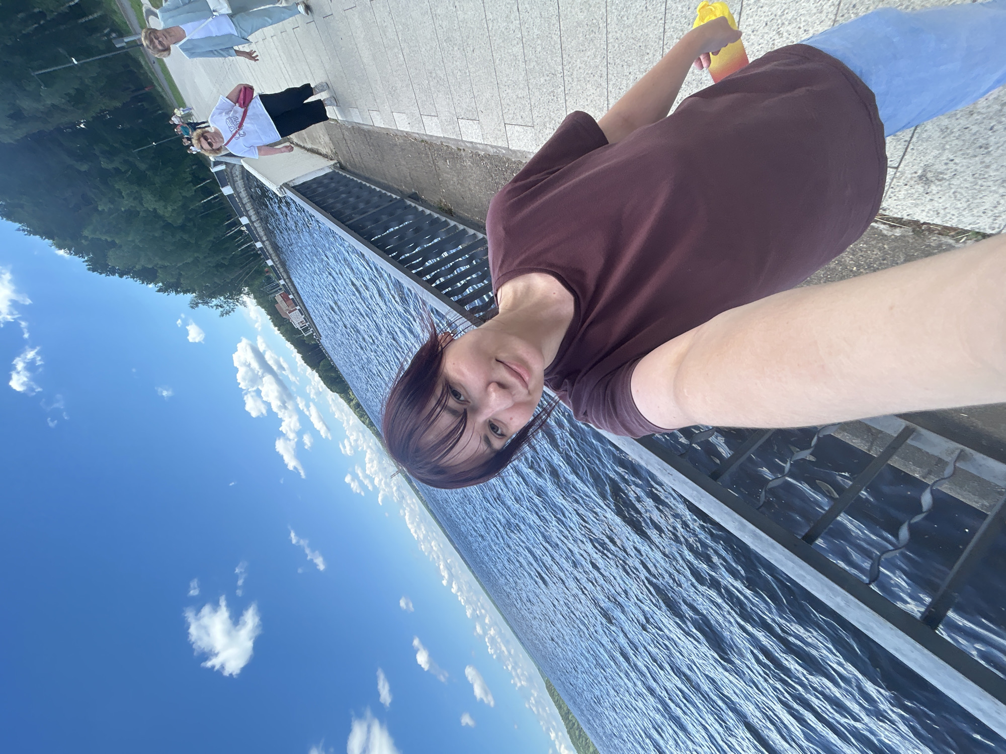 The image shows a group of people standing along a paved walkway next to a body of water. The sky is blue with scattered white clouds. In the foreground, a woman is taking a selfie, holding a yellow and orange bottle. She is wearing a maroon t-shirt and blue jeans. Behind her, other people are visible, some walking and some standing. One woman in the background is wearing a white t-shirt with text on it and a pink bag. The scene appears to be outdoors on a sunny day, possibly in the afternoon. The walkway seems to be elevated, with a railing overlooking the water. The water has gentle ripples. The background shows trees and distant structures. The overall mood seems casual and relaxed, consistent with a day out.