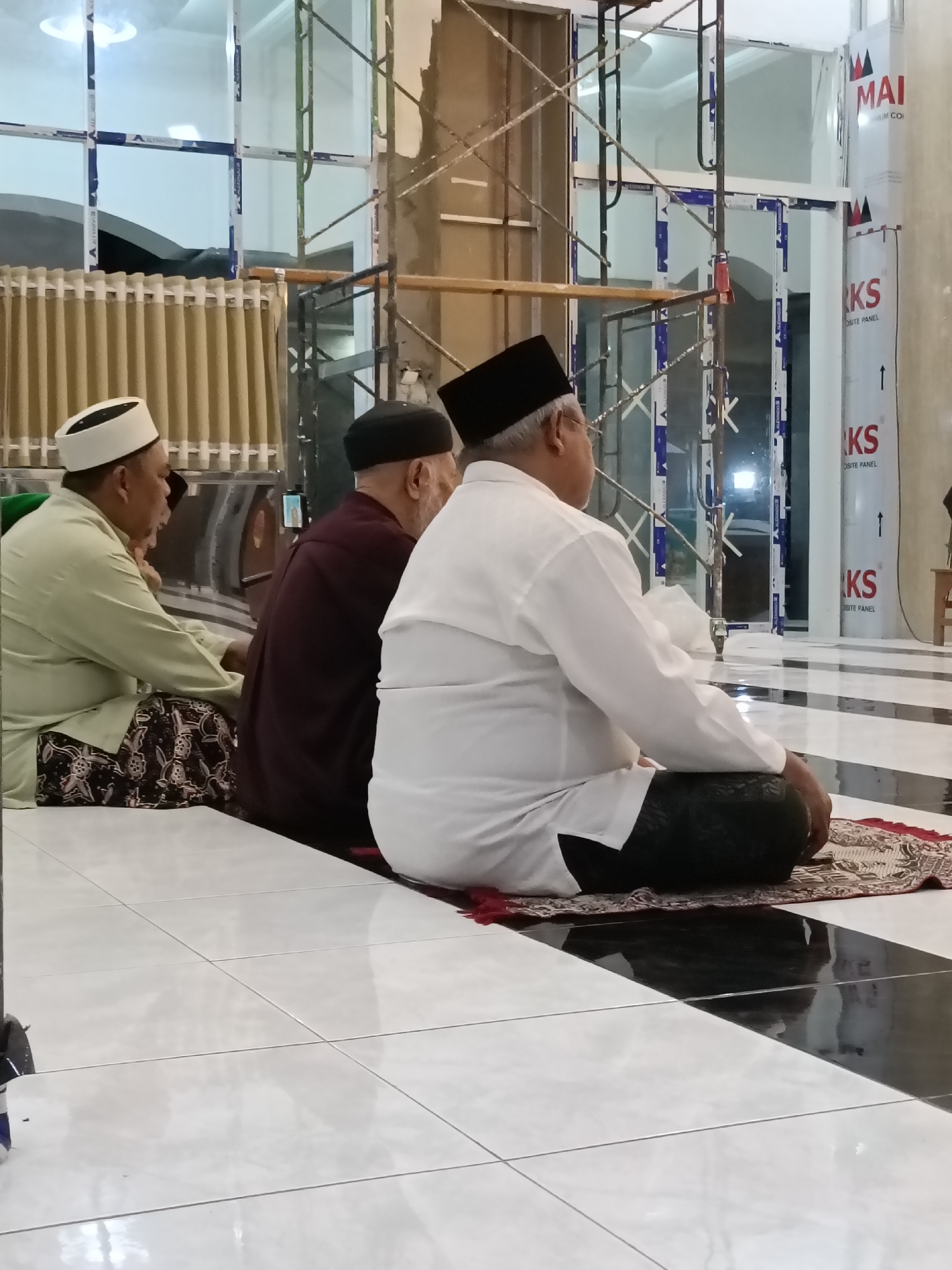 The image depicts three men in traditional attire, likely in a mosque or a prayer room in Kediri City, Indonesia. The men are seated on the floor, facing right, with the man in the foreground wearing a white shirt and black pants. He has a prayer mat beneath him. To his left, another man in a dark colored robe and a black cap is also seated. On the far left, a third man in a light green shirt and patterned sarong is seen. The environment appears to be under construction or renovation, with scaffolding visible in the background, along with large windows. The floor is tiled in a black and white checkered pattern.
