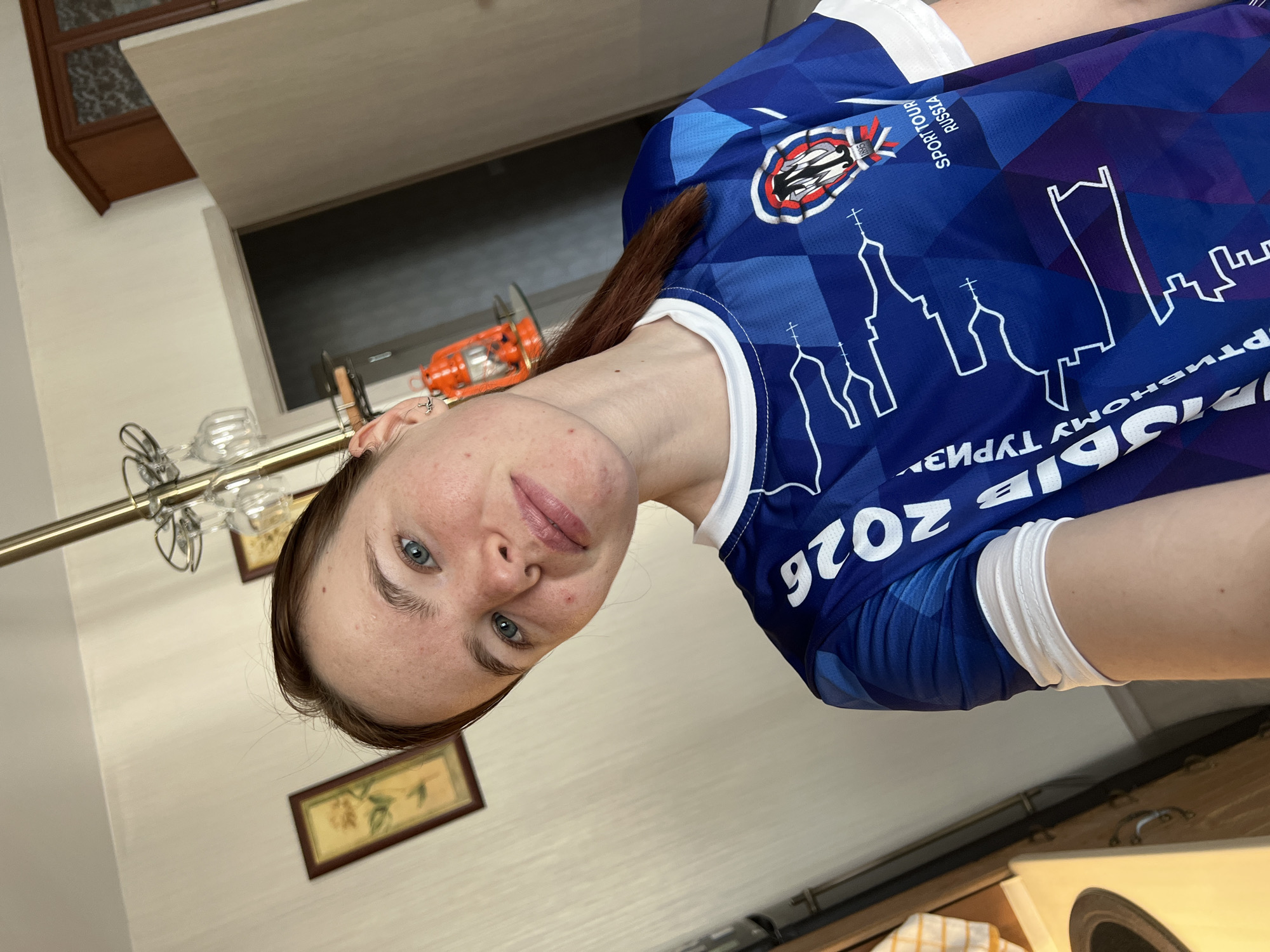 A fair-skinned woman with blue eyes is wearing a blue jersey with "RUSSIA SPORTTOUR" printed on it. The jersey also has a white skyline design of what appears to be the city of Perm, Russia, including what appear to be stylized representations of religious buildings and city structures. "2026" is also printed on the jersey. The location appears to be inside a home, evident by the presence of a chandelier and framed art on the wall. There is also a counter-top with a small speaker in the lower right corner of the image. There is no interaction evident in the image.