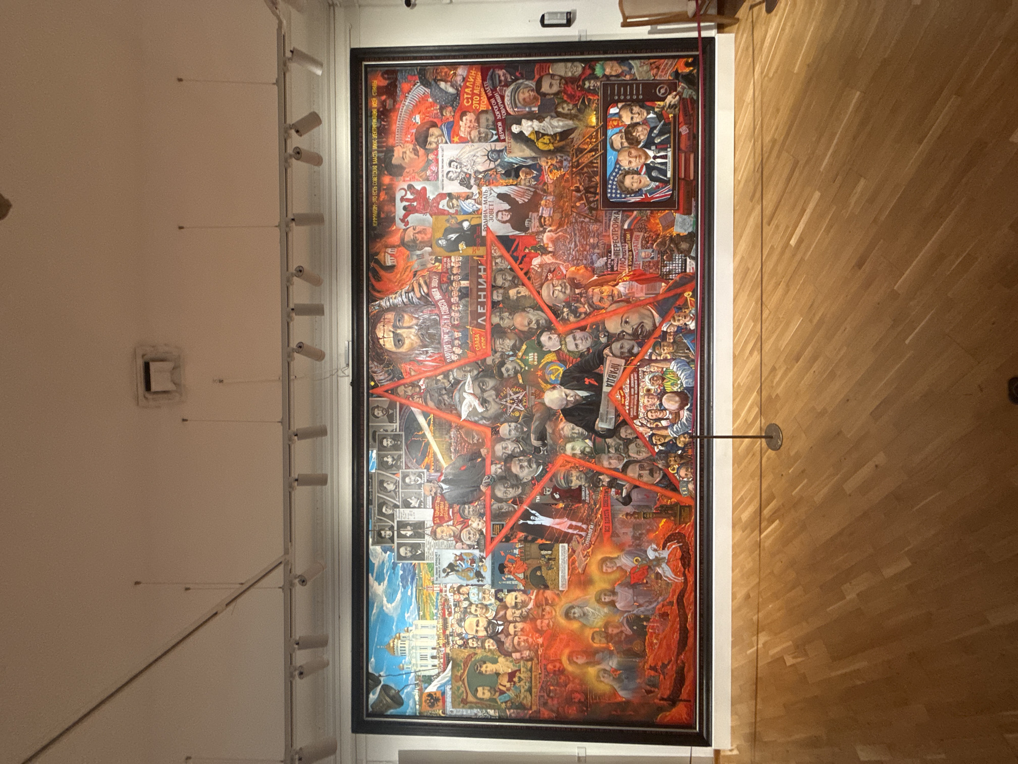 This is a wide, panoramic photograph of a large, multi-panel mural displayed on a white wall in a museum setting. The mural is framed in black and depicts a collage of historical figures and scenes, primarily related to Russian history and Soviet ideology, overlaid with a prominent red star.
The mural's content includes numerous portraits of political leaders such as Lenin and Stalin, alongside depictions of industrial scenes, agricultural imagery, and revolutionary moments. There are also visual references to the Tsarist era, with portraits of figures like Nicholas II and his family. A television screen within the mural shows portraits of Ronald Reagan and Mikhail Gorbachev. The overall aesthetic is rich and detailed, utilizing a vibrant color palette with a strong emphasis on reds and blues.
The surrounding environment consists of a light-colored ceiling with track lighting fixtures and ventilation elements. Below the mural, the floor is made of polished wooden planks laid diagonally. A thin black cord runs along the bottom of the mural, and a thin black pole with a circular base stands in front of the mural's center, likely to prevent visitors from approaching too closely. To the right of the mural, a portion of a wooden chair is visible.
This mural is located in the Novosibirsk State Art Museum in Novosibirsk, Russia. The museum is situated in the city center, on the left bank of the Ob River.
