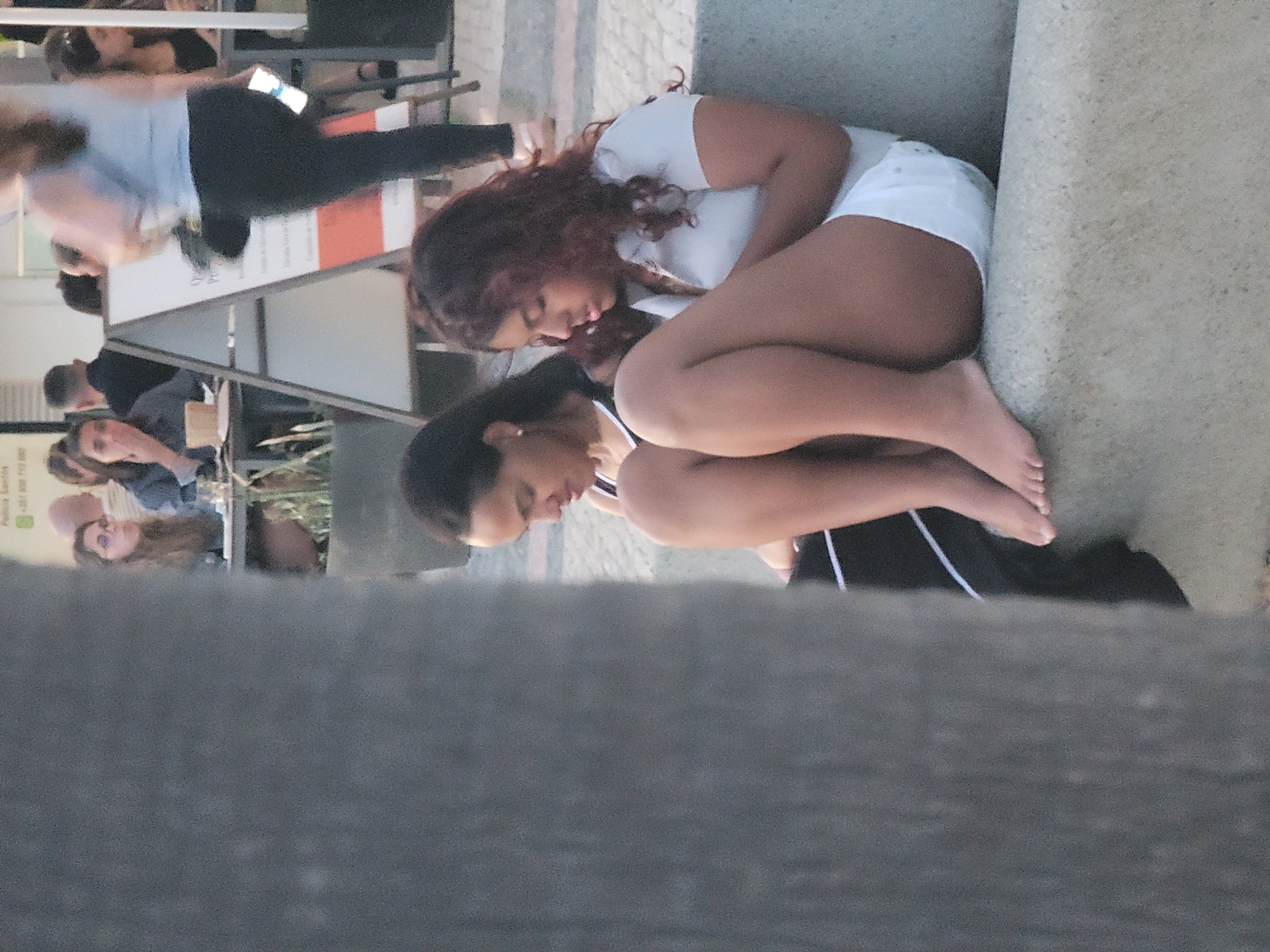 Two young women are sitting on a concrete bench outdoors. The woman on the left has dark hair and is wearing a black top with white trim. The woman on the right has reddish-brown curly hair and is wearing a white polo shirt and white shorts. Both women are barefoot. They appear to be looking down, possibly at their phones, which are not clearly visible. In the background, other people are seated at tables, suggesting a cafe or restaurant setting. A sign is visible behind the women. The scene is located in Quarteira, Portugal, near the beach. The overall lighting suggests it is daytime, likely late afternoon or early evening due to the warm tones.