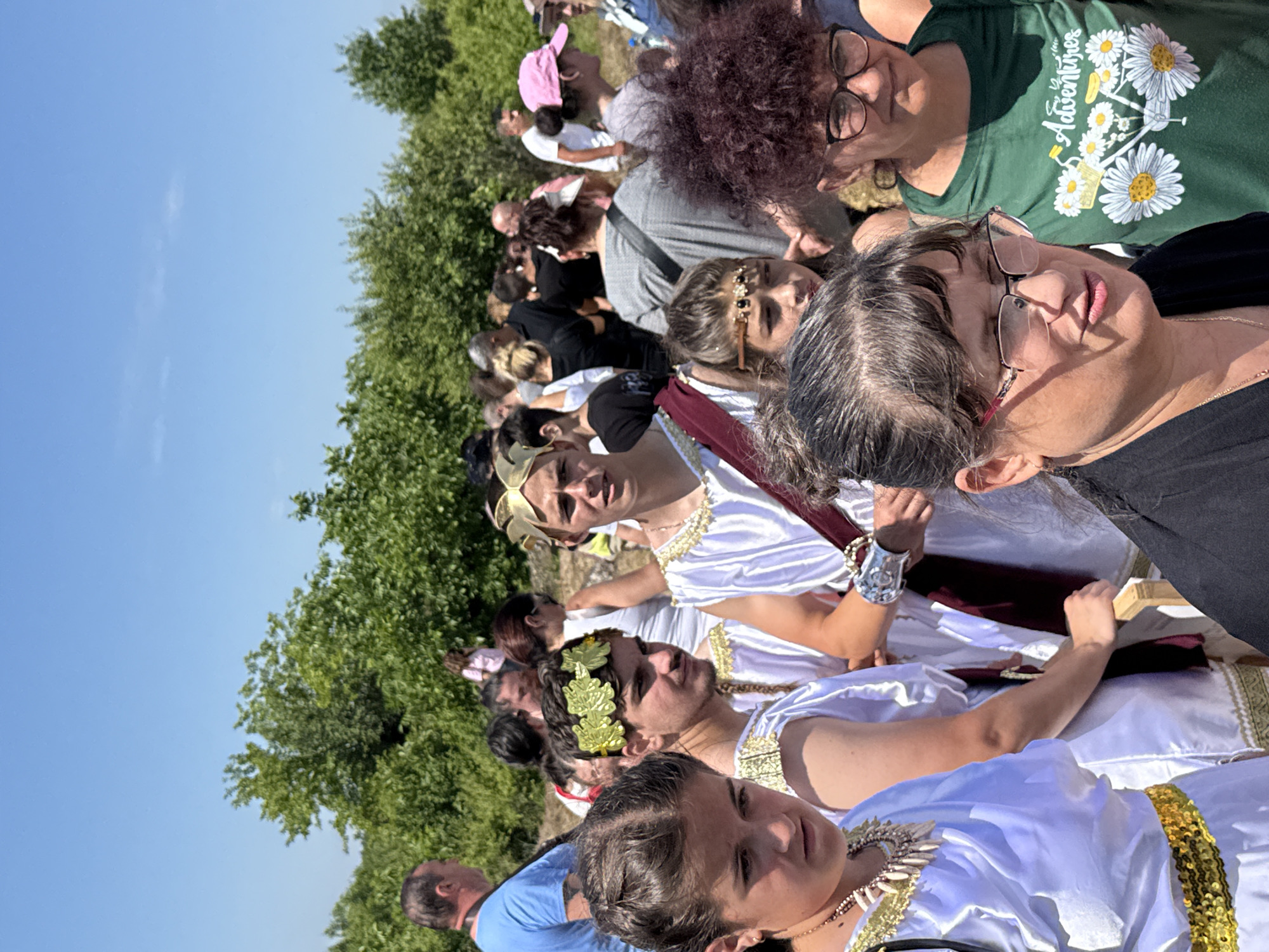 The image captures an outdoor gathering in Gigen, Bulgaria, under a clear blue sky on a bright, sunny day. The scene features a crowd of individuals, with a prominent group in the foreground dressed in attire reminiscent of ancient Roman or Greek periods, interspersed with people in modern casual clothing.
In the immediate foreground, a woman with dark hair and glasses is partially visible on the right, wearing a black top. Behind her, another woman with curly reddish-brown hair and glasses wears a dark green t-shirt adorned with white daisies and a bicycle graphic, accompanied by the word "Adventures."
To the left in the mid-ground, a young woman with dark braided hair wears a white toga-like garment with gold trim at the shoulders and a wide gold sequined belt. She has a necklace composed of white, pointed elements resembling shells or bones. Beside her, to the right, a young man with a slight mustache wears a similar white garment with gold trim and a headpiece of green, metallic-looking leaves. Further to the right and slightly behind him, another young man wears a white robe, a gold laurel-wreath style headpiece, a maroon sash draped over his shoulder, and a silver cuff on his right forearm.
Various other individuals in modern clothing are visible in the mid-ground and background, including a man in a light blue t-shirt on the far left and several people in dark tops. The background consists of dense, lush green trees and foliage under a bright, cloudless blue sky, suggesting an open, possibly rural or historical site. The ground is not clearly visible but appears to be unpaved. The overall context suggests a cultural event or historical reenactment.