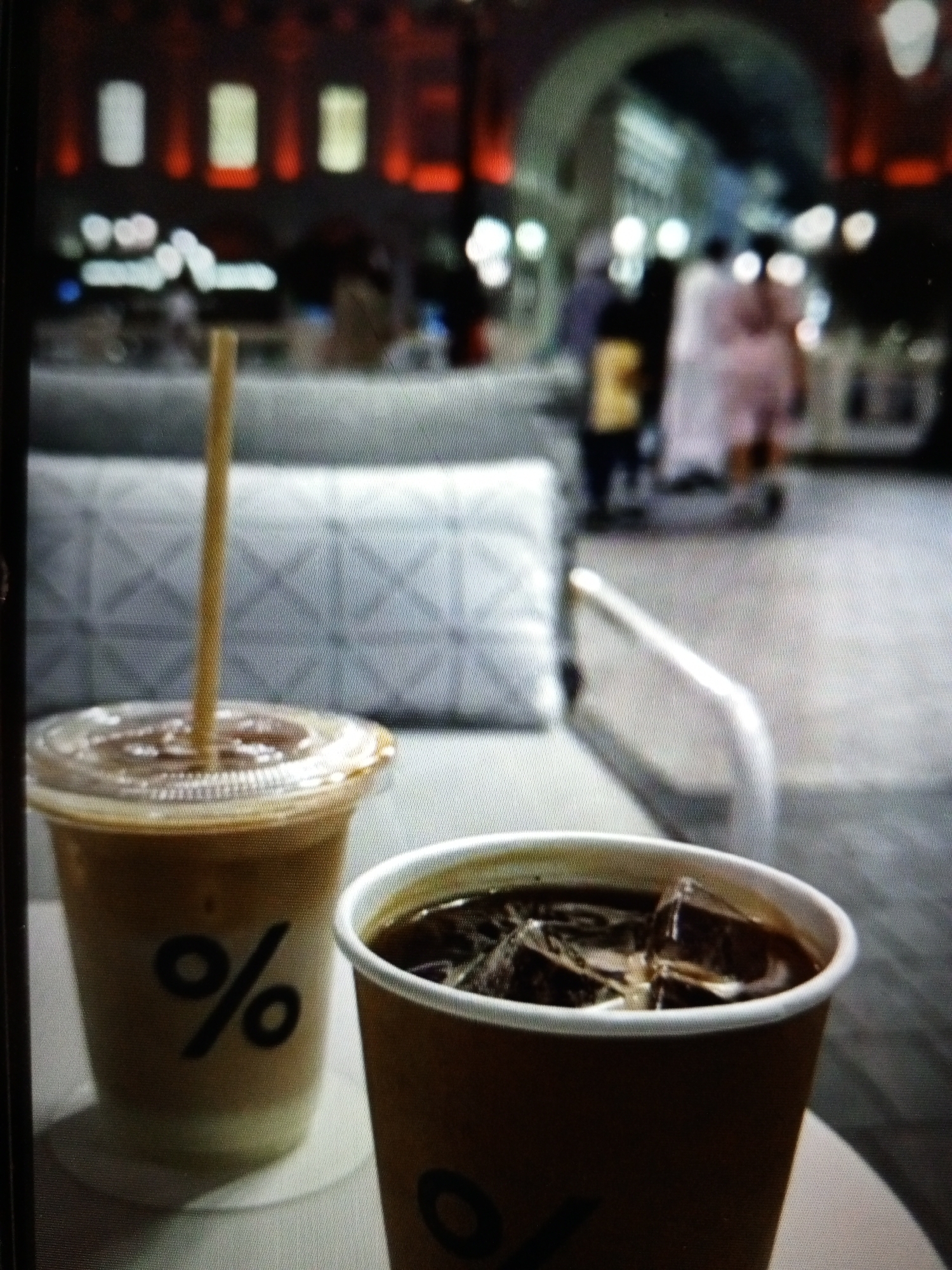 Two cold coffee drinks are placed on a table. The drink in the foreground is in a brown paper cup, filled with dark liquid and ice cubes. The second drink, to the left and slightly behind, is in a clear plastic cup with a clear lid and a brown straw. This drink has a lighter color, possibly iced coffee with milk or cream. Both cups have a black percentage sign logo. In the background, people are walking in what appears to be an outdoor market or plaza with a building featuring arched entrances and illuminated windows. The setting is a city street, potentially in Golbembal, Nigeria.