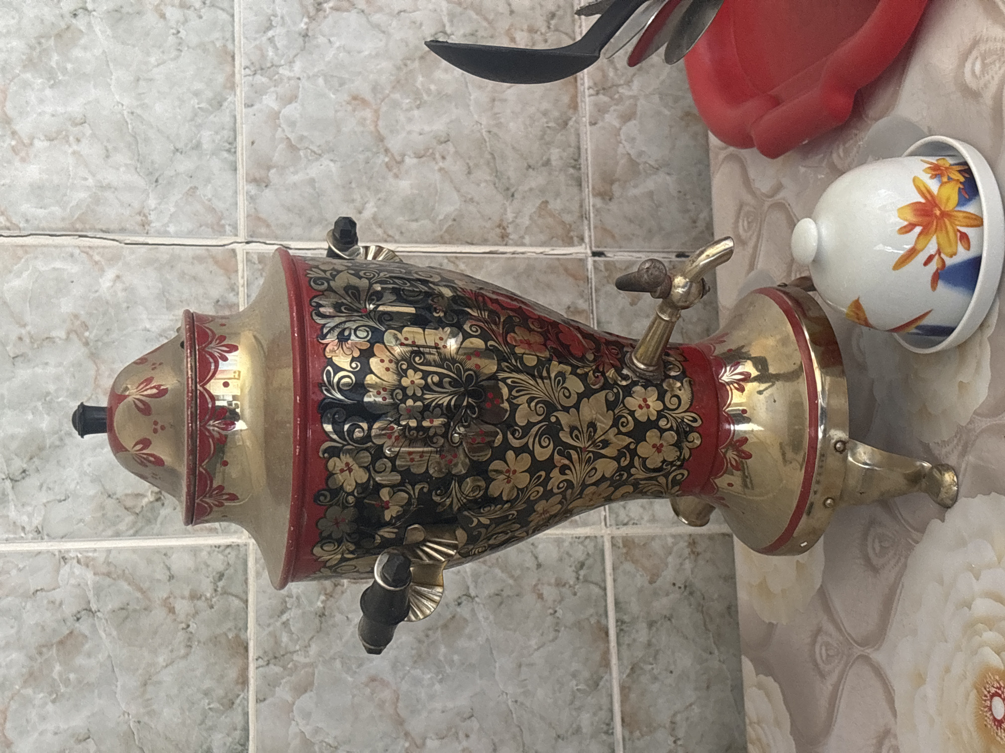 This image captures a close-up, vertically oriented view of a traditional Russian samovar, set against a tiled and wallpapered background in what appears to be a kitchen or dining area in Chaikovsky, Russia.
The central subject is an elaborately decorated samovar, likely made of brass or gold-toned metal. Its body is adorned with intricate black and gold floral and leaf patterns, accented by striking red bands near its top and base. It features a small, ornate spigot (tap) on its right side, a lid topped with a black knob, and visible handles on both sides. The samovar stands on a sturdy, decorated base with three visible legs.
To the right of the samovar, slightly behind it, a white ceramic cup is seen resting upside down on a small white saucer or lid. The cup is decorated with vibrant orange and yellow floral designs and hints of blue. Above the samovar and cup, several kitchen utensils, including a black spatula and other metallic items, are partially visible, along with a red plastic container.
The background consists of light-colored, possibly beige or gray, marbled wall tiles with dark grout lines, visible on the left and upper sections of the image. To the right, behind the samovar's base and the cup, the wall is covered with wallpaper featuring large, soft white and cream floral patterns.
The scene depicts a domestic setting, suggesting a space for preparing and serving tea, a traditional practice often associated with samovars in Russia. The lighting is bright and even, indicating either well-lit indoor conditions or daytime. There is no visible text in the image. The overall impression highlights traditional craftsmanship and a cozy atmosphere.