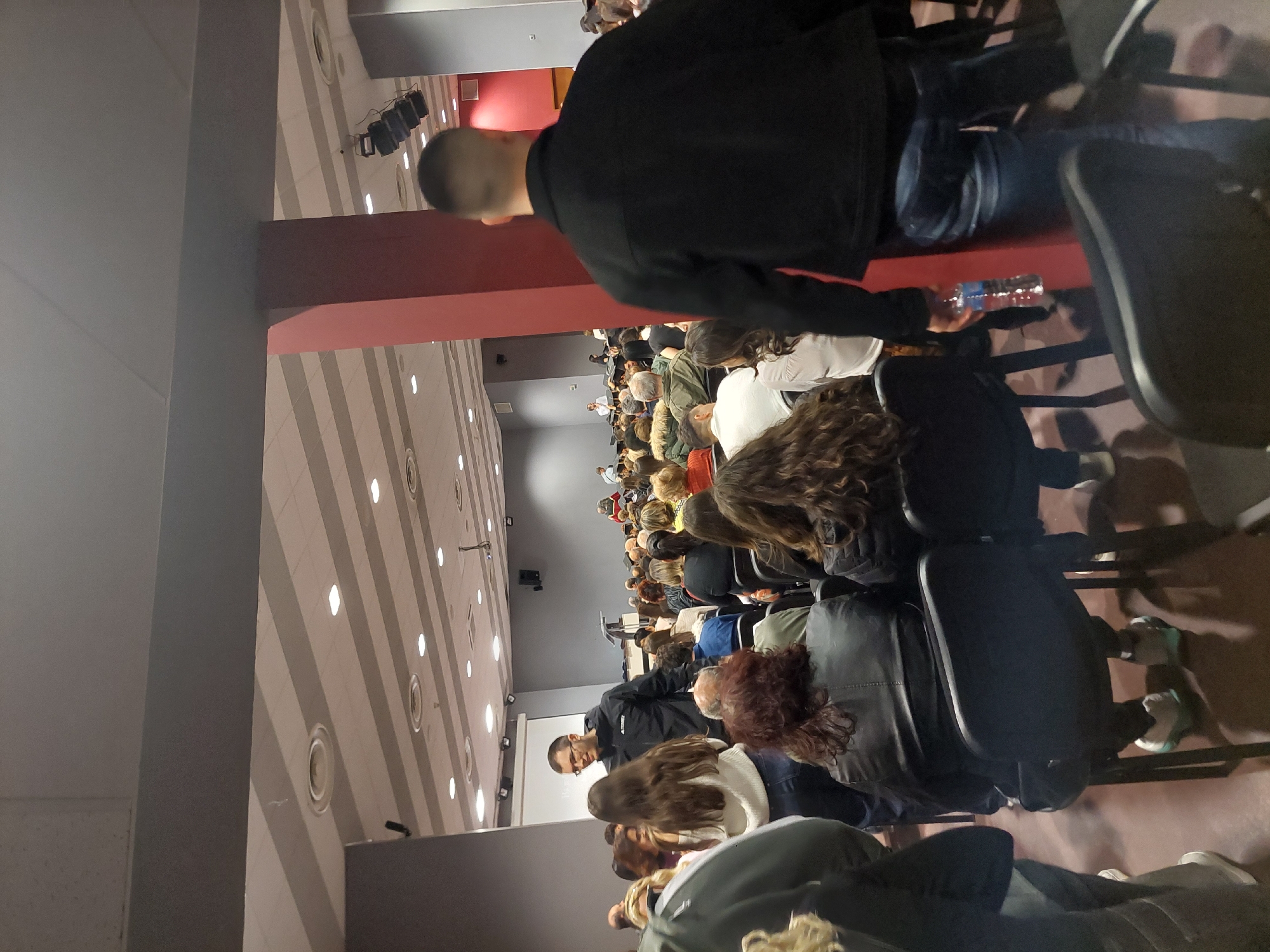 An indoor auditorium in Plovdiv, Bulgaria is filled with people seated in rows of black chairs. Most are facing forward toward a screen, presumed to be part of a presentation. The environment is well-lit, with striped ceiling lights. There is a red architectural beam along the ceiling of the space. An individual wearing a black jacket is walking along the right of the frame, carrying a water bottle. The audience is diverse.