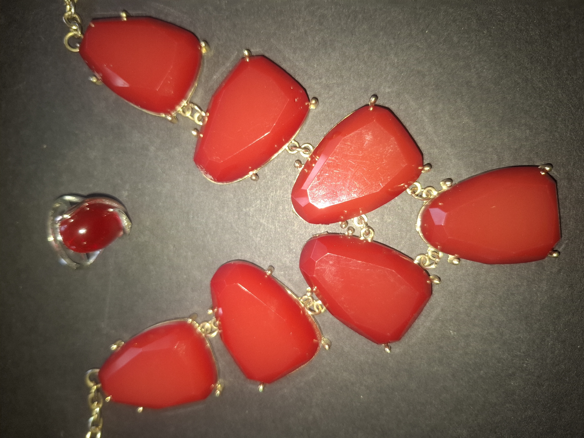 A red gemstone necklace and ring are displayed against a dark, textured background. The necklace is gold-toned and features a cascade of seven faceted, tear-drop shaped red gemstones. A matching ring with a single red gemstone is positioned above the necklace. The lighting creates highlights and shadows on the gemstones, emphasizing their polished surfaces. The scene appears to be a product shot, possibly for e-commerce. There is no information available to determine its location in Almaty, Kazakhstan.