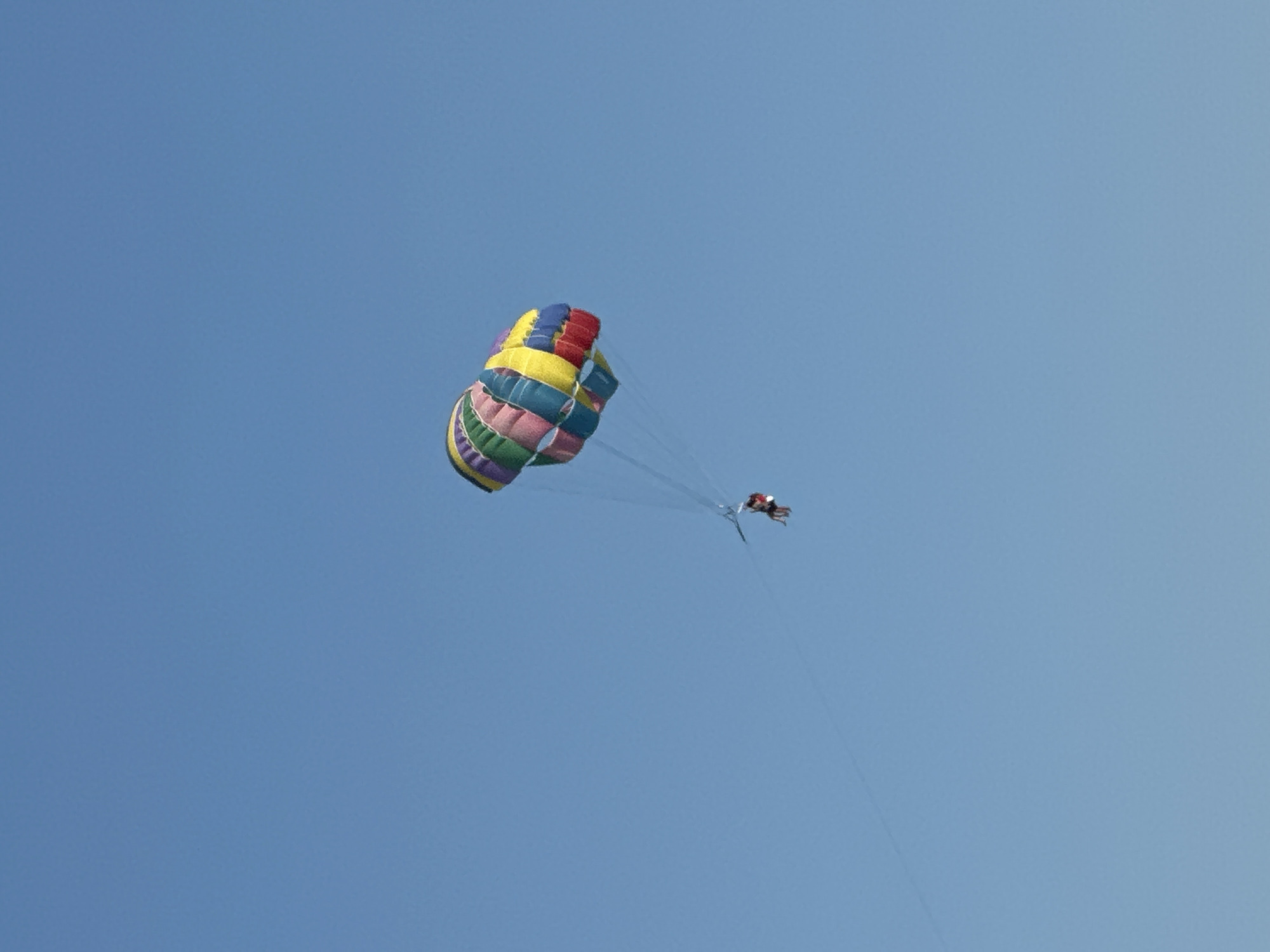 This image shows a person parasailing in the sky above Boreti, Montenegro. The parasail is colorful, with stripes of yellow, blue, green, purple, and red. Two people are visible, one in the parasail and one in the water below, possibly being pulled by a boat. The sky is clear and blue.