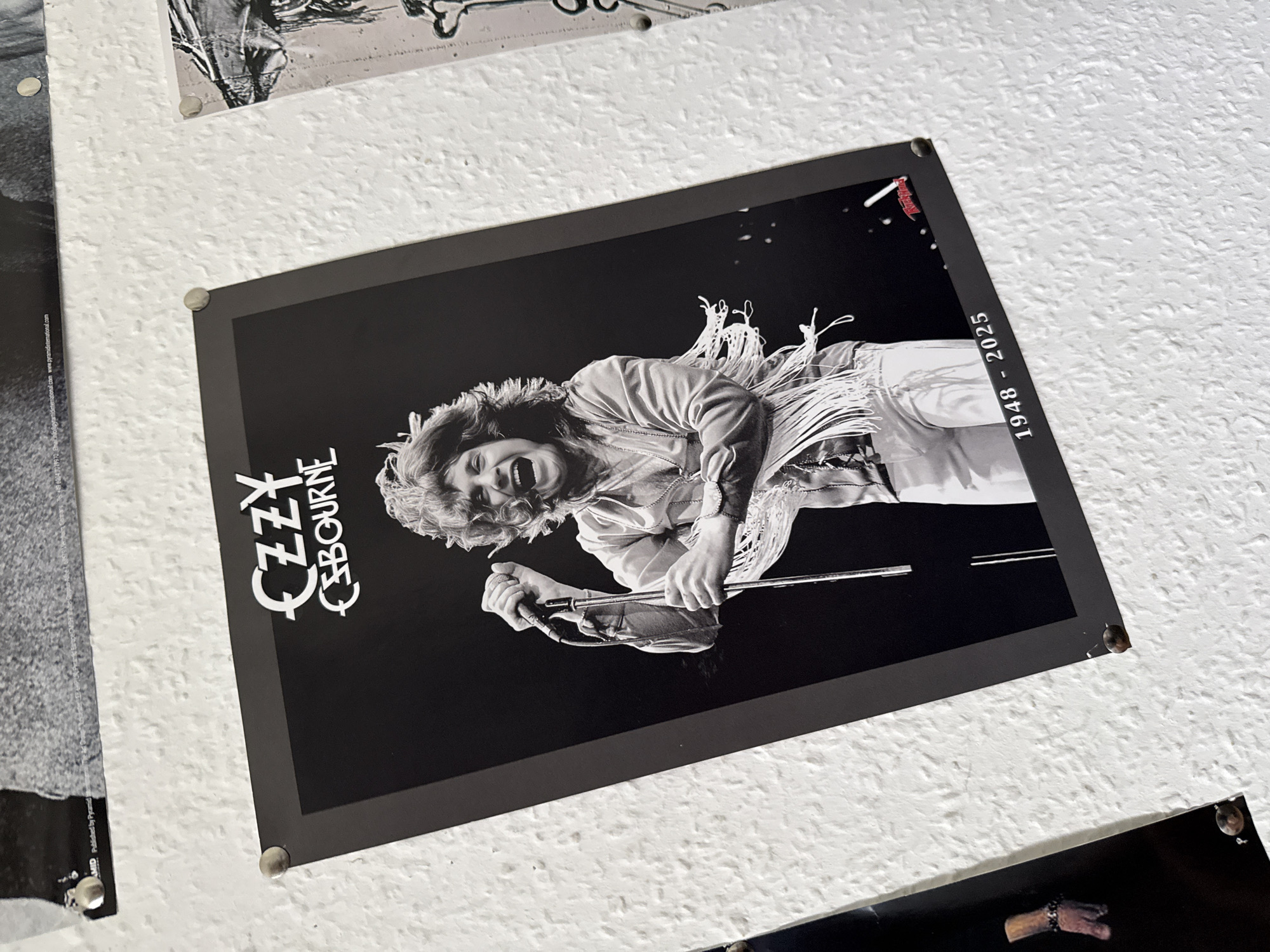 The image shows a black and white poster of Ozzy Osbourne performing on stage, with the years "1948 - 2025" printed below him. The poster is attached to a textured white wall with thumbtacks, typical of a bedroom or dorm room. The poster is slightly tilted, and the lighting suggests it is indoors. There are other posters partially visible in the background, also attached to the wall. The poster depicts Ozzy Osbourne with his mouth open, holding a microphone and wearing a fringed shirt. The poster itself has a dark border and the text "OZZY OSBOURNE" is prominently displayed at the top. There is a red logo in the bottom right corner of the poster. The overall impression is of a fan's personal space decorated with rock memorabilia. This scene is located in Bad Sachsa, Germany.