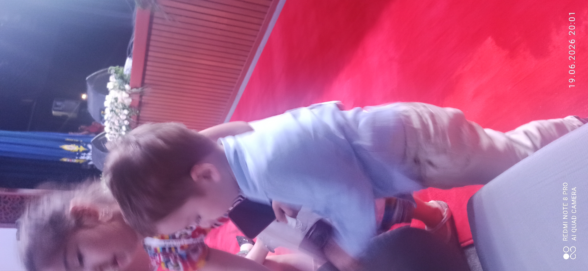 In an indoor setting with a red carpeted floor and wooden paneled walls, a young boy wearing a light blue collared shirt and tan pants is captured mid-stride. He is holding a dark rectangular object, possibly a phone, in his left hand. To his left, another child, a girl, is partially visible, wearing a colorful patterned top. In the background, a blue curtain with a golden emblem hangs, and a stage decorated with white flowers is present. The scene appears to be illuminated by artificial lighting, creating a soft glow. A date and time stamp, "19.06.2026 20:01", is present in the bottom right corner.