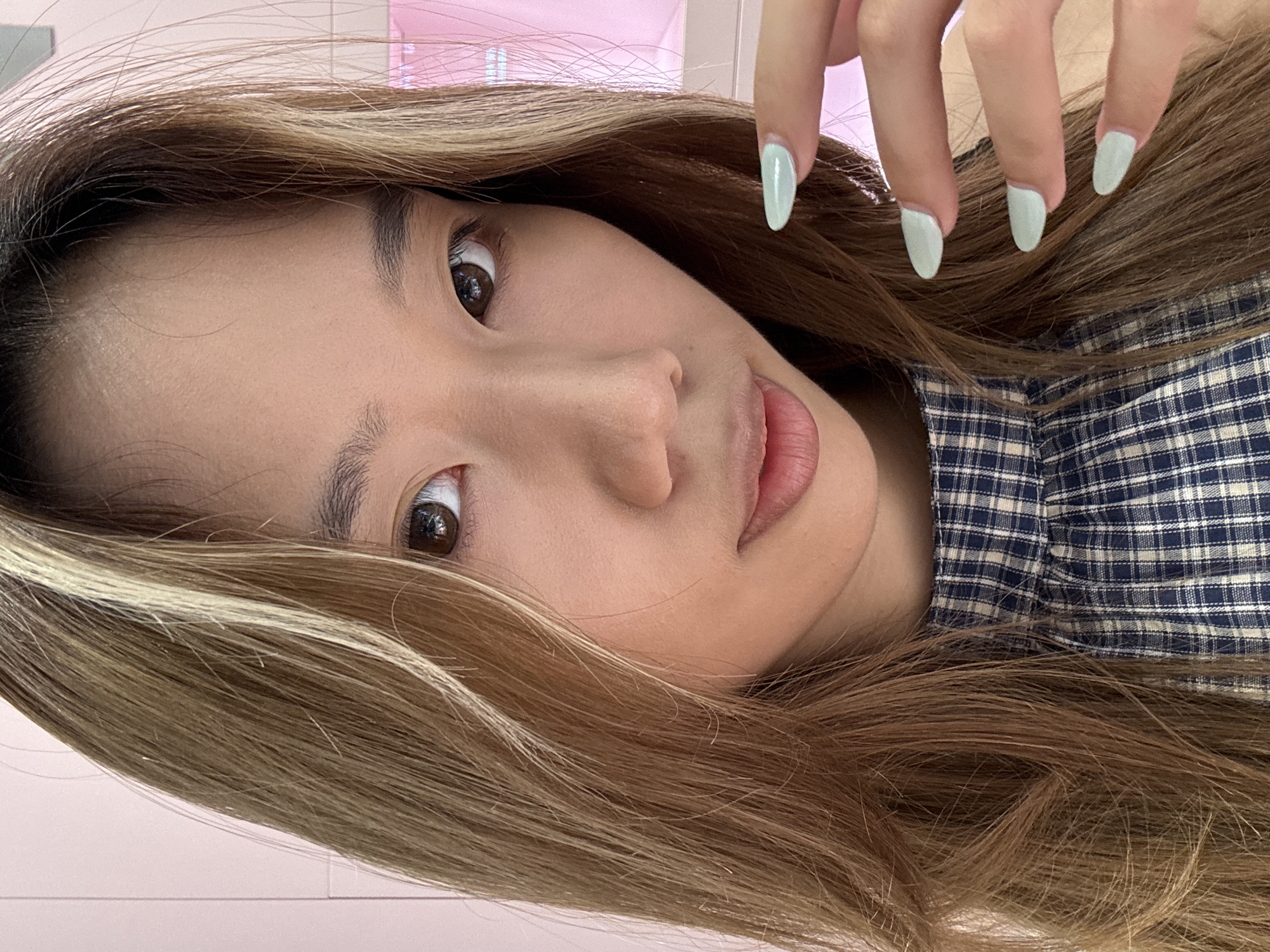 The image displays a close-up, angled portrait of a young person, likely female, against a soft, blurred pink background, suggesting an indoor setting in Liangjiang, China. The subject's head is tilted significantly to the left, and they are looking directly at the camera with a neutral and calm expression.
The individual has light brown eyes with dark pupils and appears to have smooth skin. Their lips are full and slightly parted. Long, wavy brown hair with distinctive lighter blonde or caramel highlights frames the face and extends throughout the visible portion of the image.
In the upper right corner, four fingers of one hand are visible, resting near the subject's hair. The nails are long, almond-shaped, and painted a light mint green or pastel blue-green color. The subject is wearing a blue and white plaid shirt, which is visible on the right side of the frame, covering their shoulder and chest. The lighting is soft and even. No text is visible within the image.