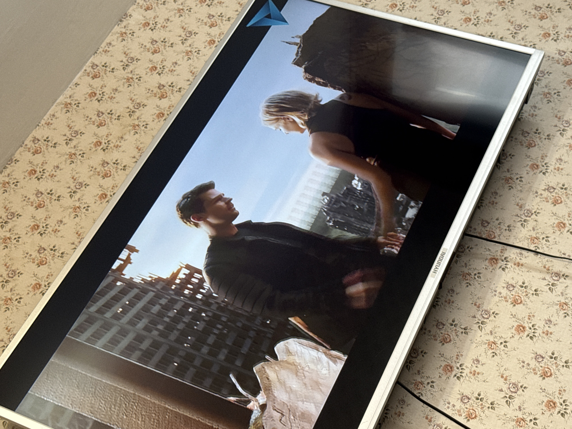 A silver-framed Hyundai monitor displays a scene of a man and woman. The man has short brown hair, and is wearing a black shirt and jacket. The woman has short blonde hair, and is wearing a black shirt. They are standing in front of a ruined city, with broken buildings. The monitor is on a surface with a floral pattern. It is not possible to identify the location in Pushkino, Russia, from the image.