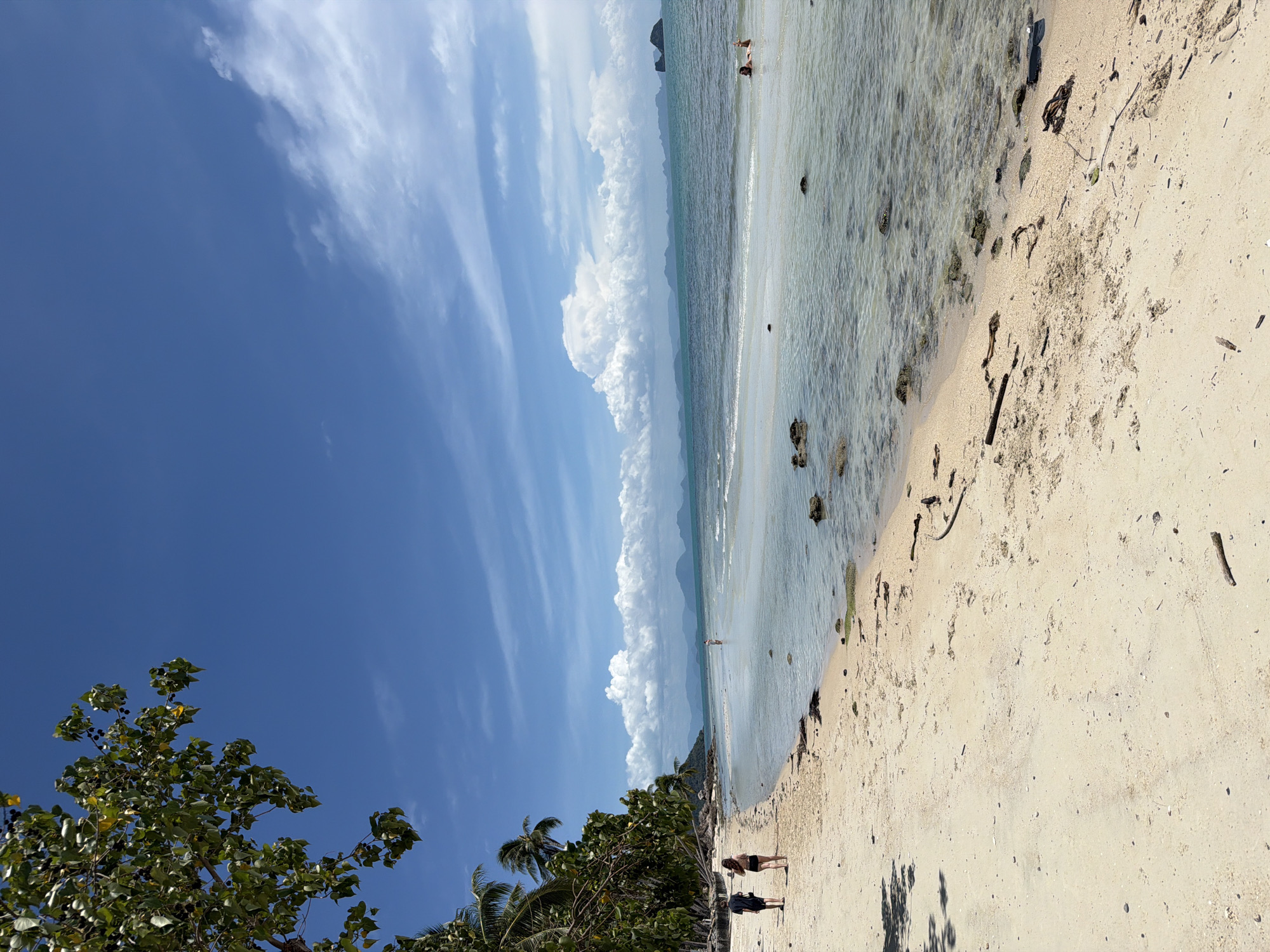 Baan Taling Ngam, Thailand: A bright beach scene with a blue sky and scattered clouds. The beach is light-colored sand, with debris like twigs and leaves. The ocean is shallow, with rocks visible in the clear water. Two people stand on the sand. Trees and vegetation line the edge of the beach. An island appears on the horizon.