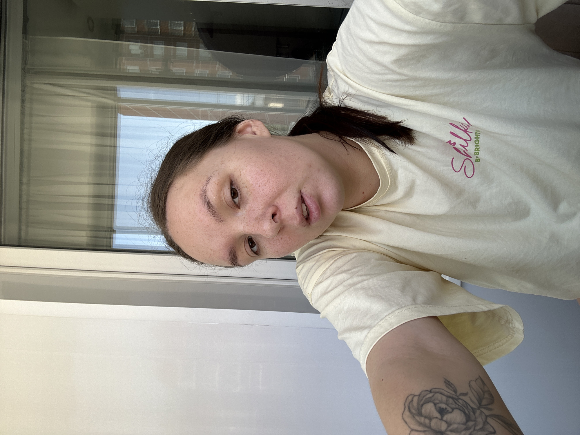 A woman is indoors, seemingly in Chaikovsky, Russia, evidenced by the building seen through the window. She is wearing a light yellow t-shirt with a pink logo and has dark hair pulled back into a low ponytail. Her skin has freckles, and she has a tattoo on her forearm. The background consists of a window reflecting a brick building. The area near the woman appears to be a white wall or door.