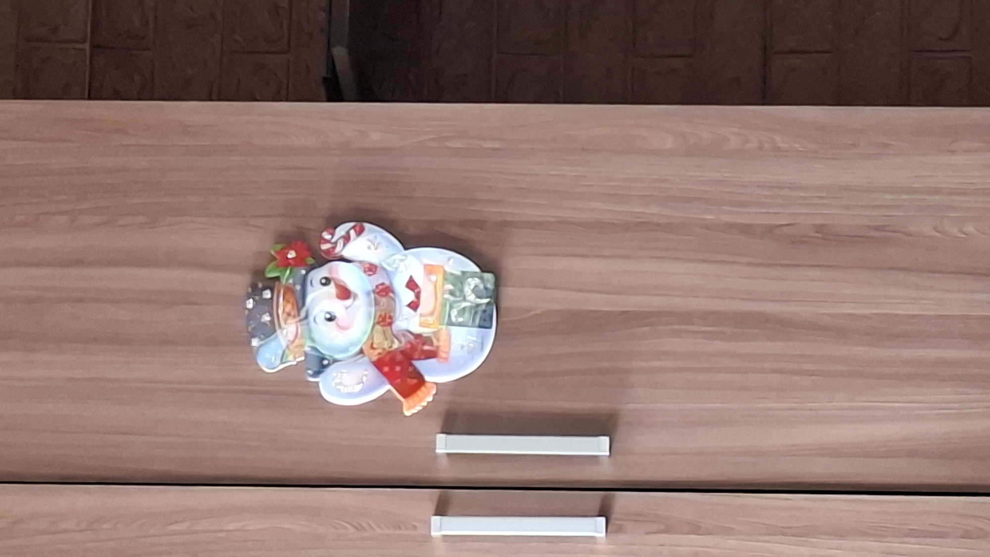 The image shows a close-up of a wooden closet door with a festive snowman decoration adhered to it. The snowman is cartoonish and appears to be made of thick paper or cardboard, with a blue hat, a red scarf, and holding a candy cane. It is positioned slightly off-center to the right. On the left side of the closet door, two white, rectangular handles are visible. The wood grain of the door is prominent. To the right of the closet door, a section of a dark brick wall is visible. There is no indication of the city of Novoilinsky, Russia, in the image.