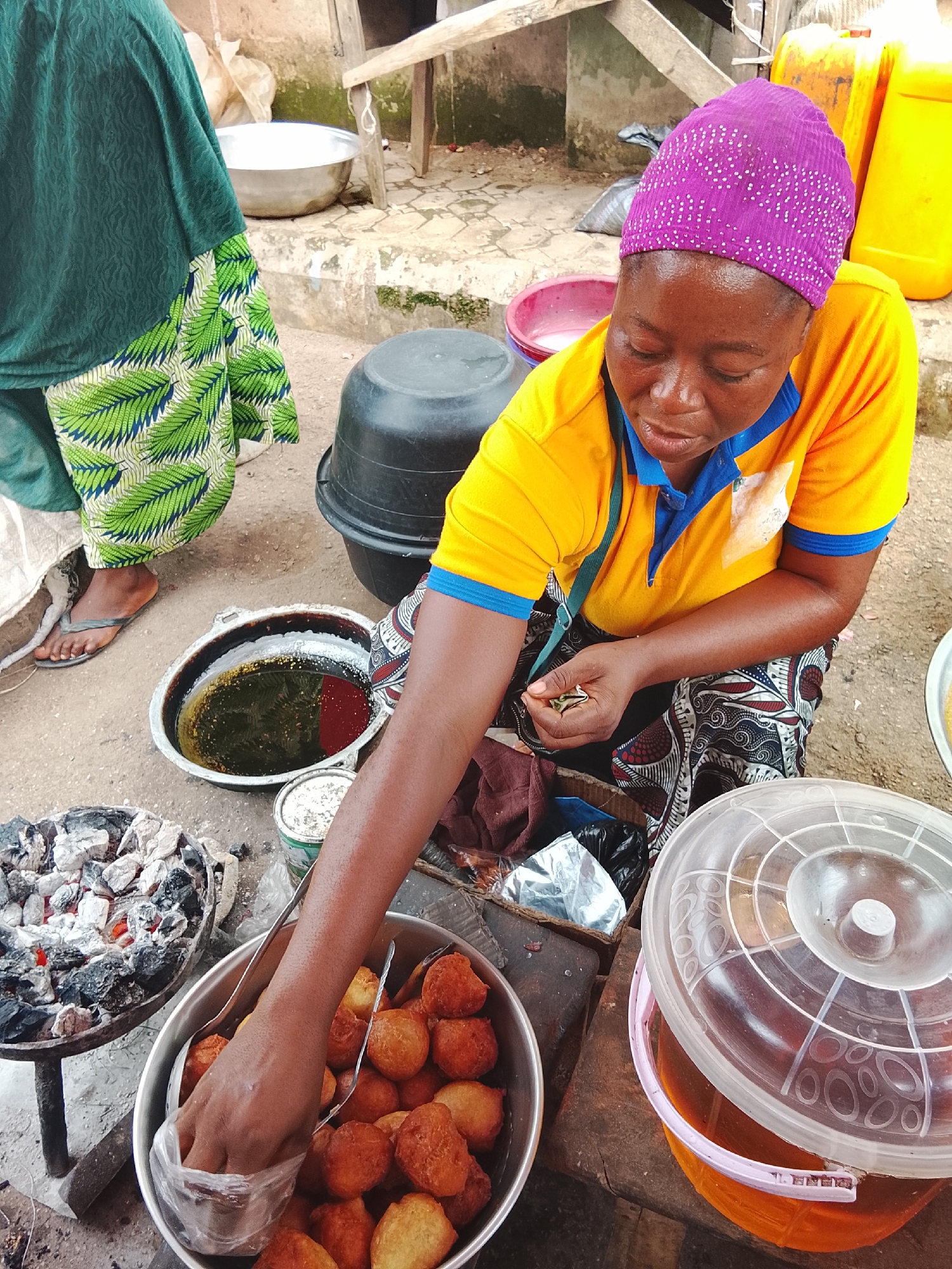 This is a medium shot of a woman, wearing a yellow t-shirt with blue trim and a purple headscarf with white polka dots, kneeling at a street-side food stall in Akwanga, Nigeria. Her dark skin is glistening with sweat. She is holding a small, golden-brown fritter in her left hand, seemingly preparing to fry it or place it in a bag. In front of her is a large metal bowl filled with numerous more fritters, some of which are being handled by metal tongs. To her left, a shallow, oval metal pan contains hot oil with some fried fritters floating in it, adjacent to a small pile of glowing charcoal embers. Further to the left, a person wearing a green headscarf and a green and white patterned skirt is partially visible. To the woman's right is a large orange plastic container with a clear plastic lid, filled with a reddish-brown liquid, likely oil. The ground is a mix of dirt and concrete. In the background, a wooden structure is visible, along with large yellow plastic containers and black buckets. The overall scene suggests a bustling food vending operation.