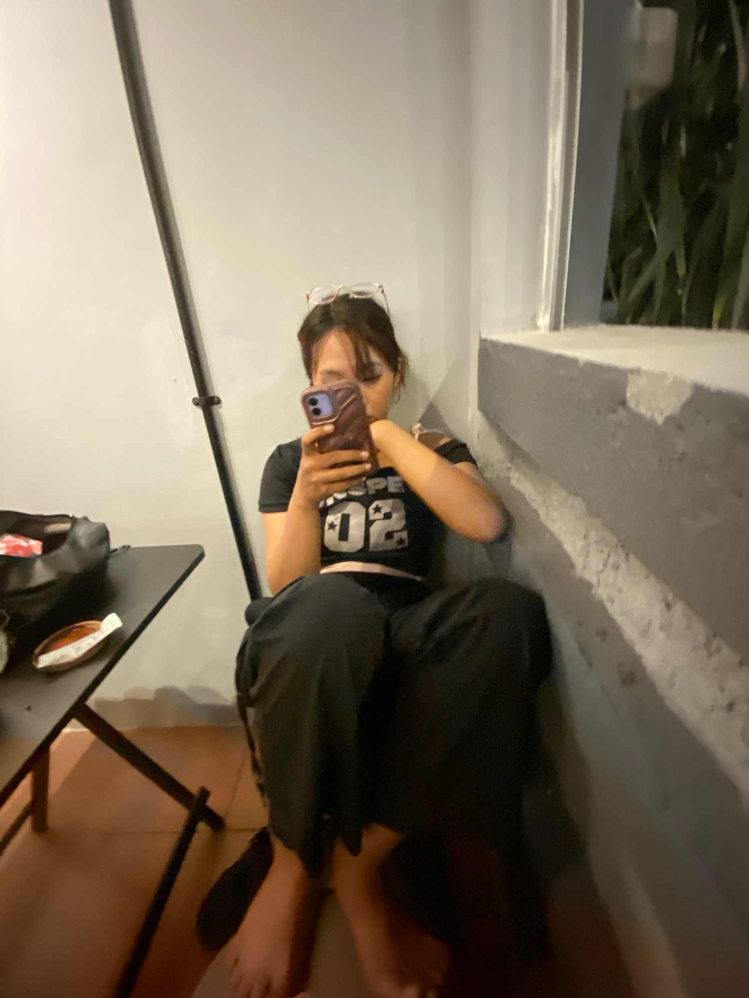 A young woman with dark hair, wearing glasses, is sitting on the floor with her back to a light gray wall. She is looking down at her phone, which she holds in her right hand. Her left hand is resting on her thigh. She is wearing a black crop top with a white number "02" and a star graphic on it, and black pants. To her left is a black folding table with a black bag on it. The bag contains some items wrapped in red and white. On the table is a small oval dish with a white wrapper. The floor is tiled in a reddish-brown color. Behind her and to her right is a window with a concrete ledge, and beyond that, some green foliage can be seen. A thin, dark gray pole is visible vertically to her left, running from floor to ceiling. The scene appears to be an outdoor seating area or balcony, possibly in the city of Klurak, Indonesia, given the context. The lighting is dim, suggesting it is either dusk or nighttime.