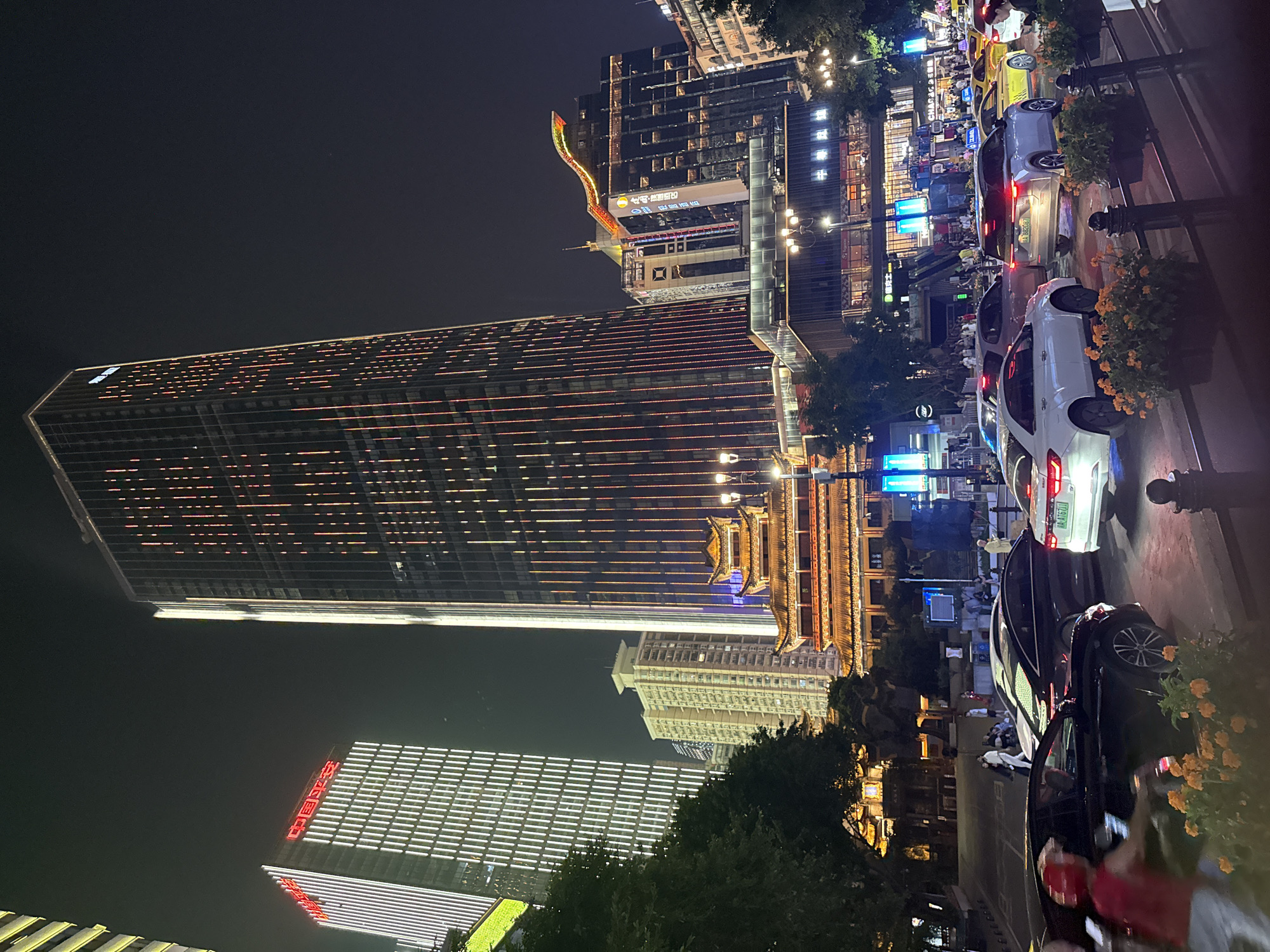 This image captures a nighttime street scene in Yuzhong District, Chongqing, China. A tall skyscraper dominates the background, its facade adorned with vertical lines of red and white lights. To its left, another skyscraper is illuminated with vertical white stripes, and red Chinese characters are visible on its upper section. In the midground, a traditional multi-tiered building with glowing orange accents and a pagoda-style roof sits between the skyscrapers. The street in the foreground is filled with traffic, including sedans and taxis, their headlights and taillights illuminating the night. Several people are walking on the sidewalk, and a person in a red vest and helmet is partially visible in the lower left corner, appearing to be in motion. Streetlights and trees with dark foliage line the street, contributing to the urban nighttime ambiance.