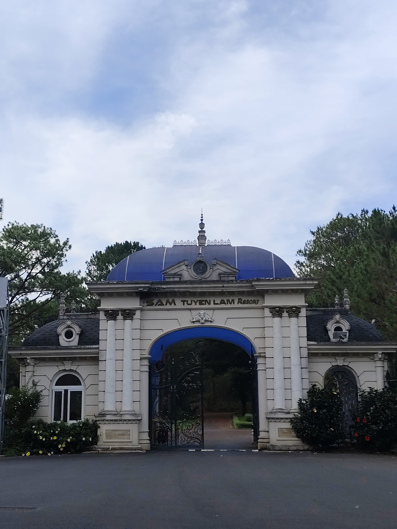 The image shows the entrance gate to the Sam Tuyen Lam Resort in Da Lat, Vietnam. The gate is a grand, classical structure with white pillars, a blue-domed roof, and ornate ironwork. The name of the resort, "SAM TUYEN LAM Resort," is inscribed above the archway. The gate is flanked by smaller structures with arched windows and dark roofs, surrounded by shrubbery with some yellow and red flowers. The sky above is overcast with clouds. The foreground is a paved road leading through the gate. Trees are visible behind the gate and on either side of the structure.