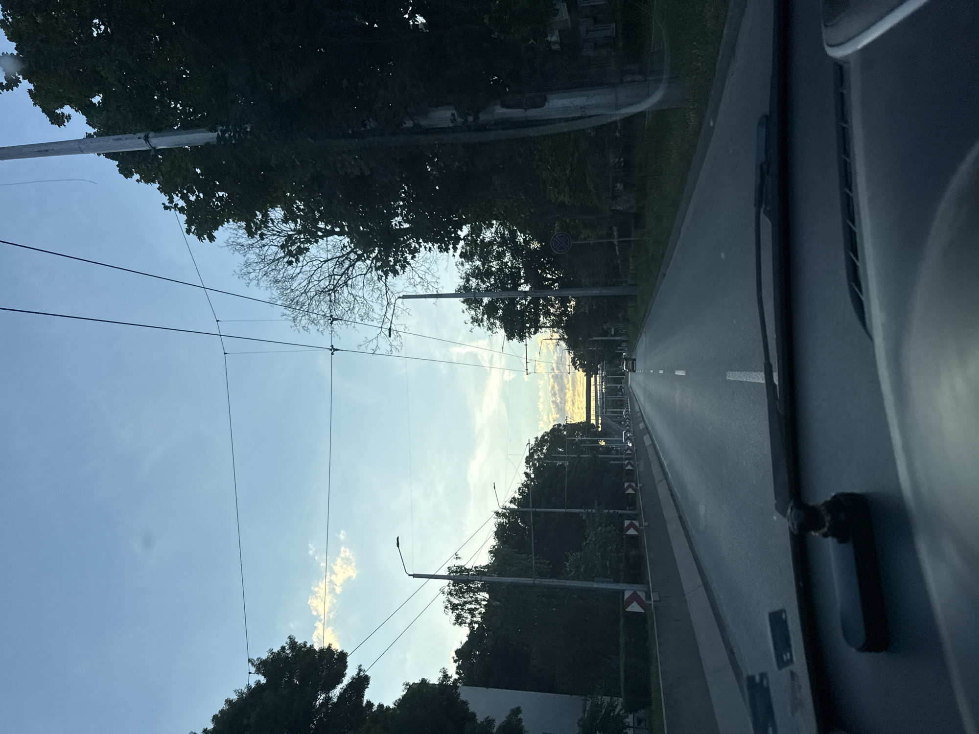 The image depicts a view from inside a vehicle traveling on a road in Varna, Bulgaria. The road appears to be a multi-lane avenue lined with trees and streetlights, with overhead power lines suggesting the presence of public transportation, likely trams or trolleybuses. In the distance, the sky is a pale blue with some scattered clouds, hinting at either early morning or late evening. The road surface is asphalt, with white lane markings visible. The vehicle's dashboard is partially visible in the foreground, with a windshield wiper and a small decorative object on the dashboard. The scene appears calm and uneventful, with only a few other vehicles visible on the road.
