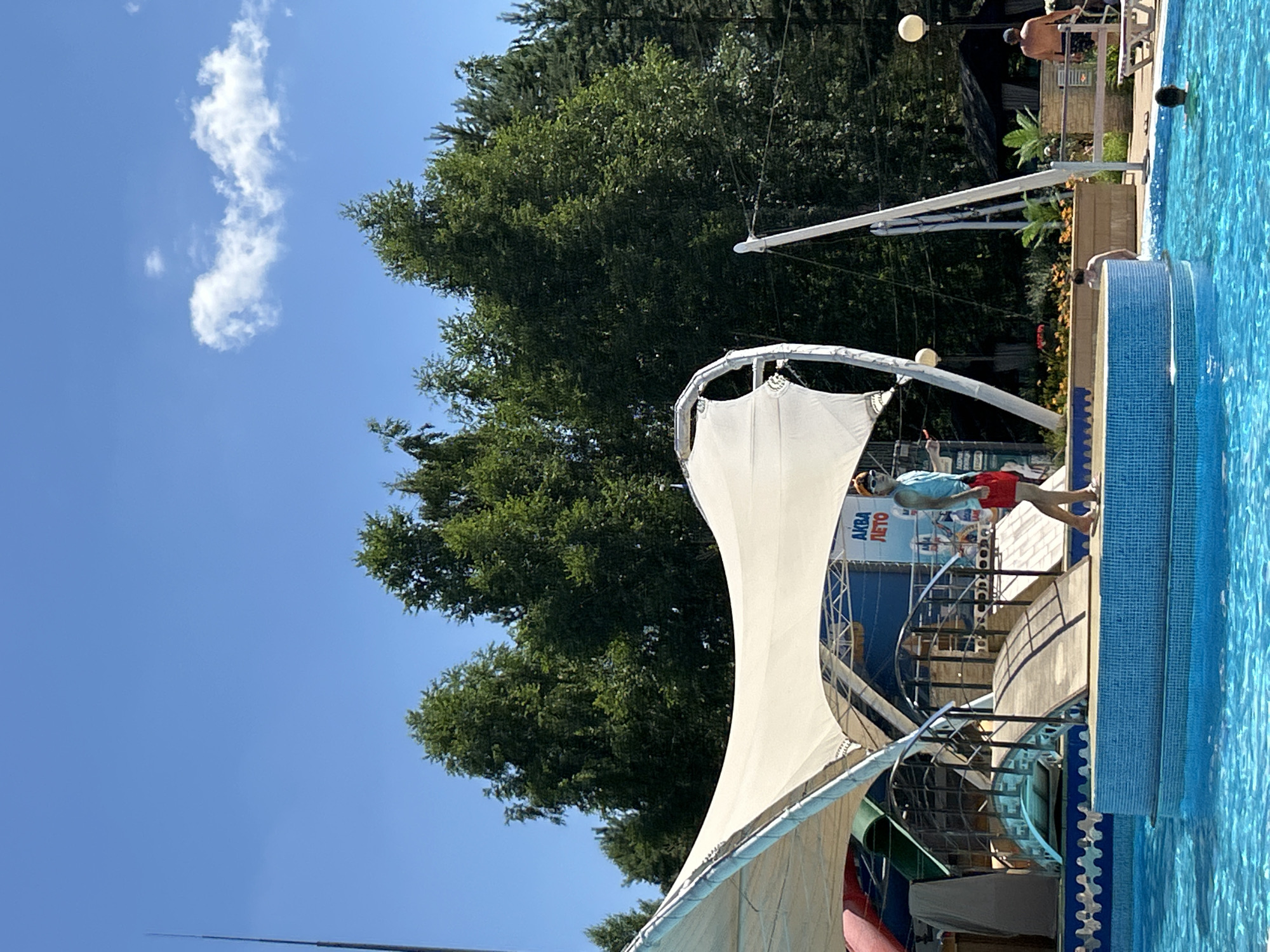 The image depicts an outdoor swimming pool area on a clear, sunny day. In the foreground, a vibrant blue pool with rippling water occupies the lower half of the frame. A tiled edge surrounds the pool, upon which a young person wearing a light blue tank top and red shorts stands. This individual is looking towards the right side of the frame. Behind them, a white, tent-like structure with curved supports partially covers a walkway and some water slides. The walkway features a decorative metal railing and a wooden bridge. In the background, lush green trees form a dense forest, with a bright blue sky overhead dotted with a few wispy clouds. A tall, thin pole with a pointed top extends vertically on the far left side of the image. Further in the background, there are glimpses of seating areas, awnings, and other people enjoying the facility. The signage visible on the structure behind the person reads "АКВА ЛЕТО," which translates to "Aqua Summer." This scene is consistent with an establishment like the "Aqua Summer" water park located in Togliatti, Russia, known for its outdoor recreational facilities.
